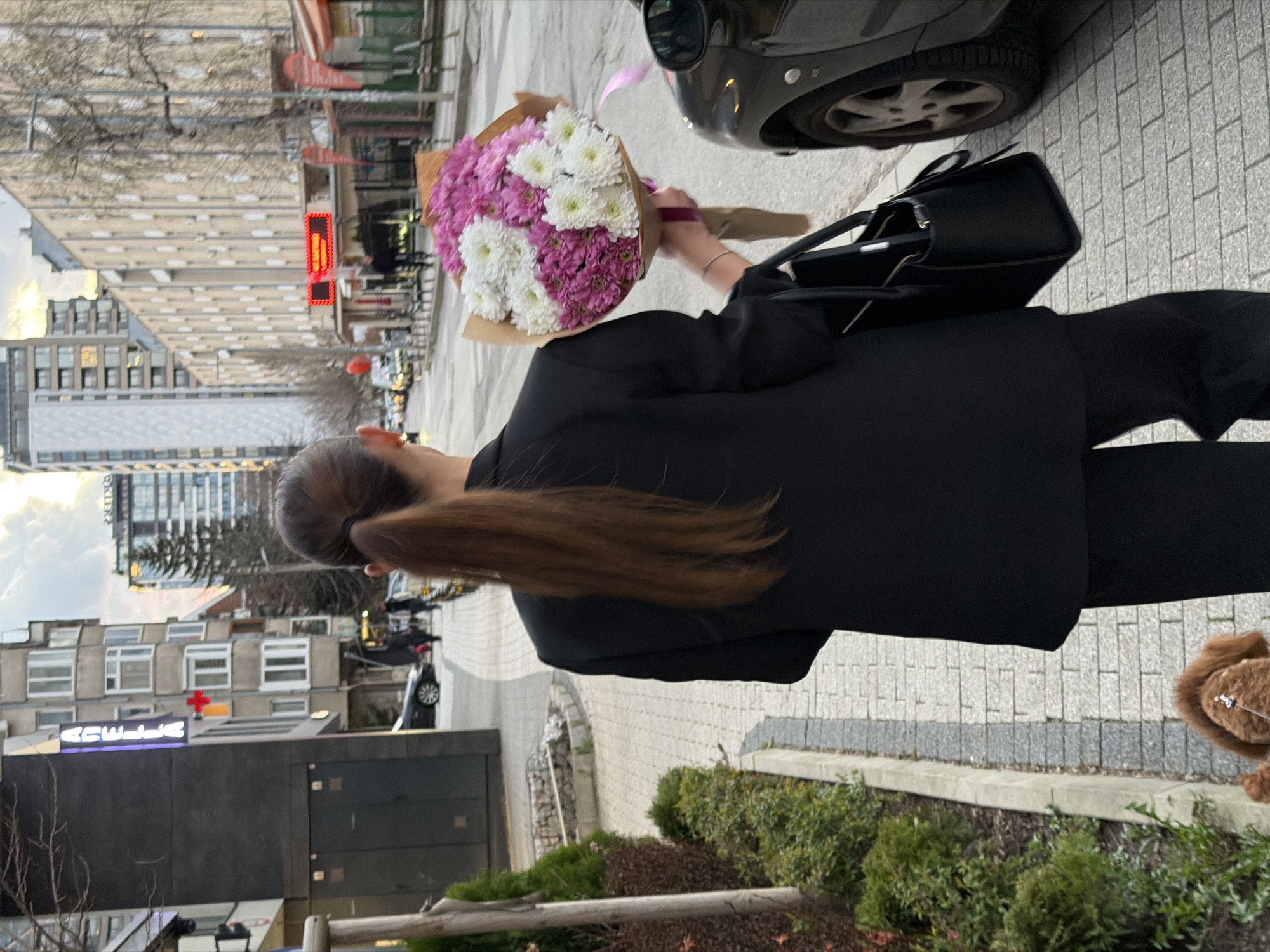 A woman in a black suit carries a bouquet of white and purple flowers and a black handbag. She is walking on a sidewalk next to a street. A car is parked nearby. The "Acibadem City Clinic Tokuda Hospital" building is visible in the background, indicating the location is likely in Sofia, Bulgaria. Shrubs grow along the sidewalk edge. The woman's hair is brown and pulled back in a ponytail. A small dog (possibly a poodle) is also visible. The streetscape shows urban buildings of various heights, typical of a city environment. The weather is overcast.