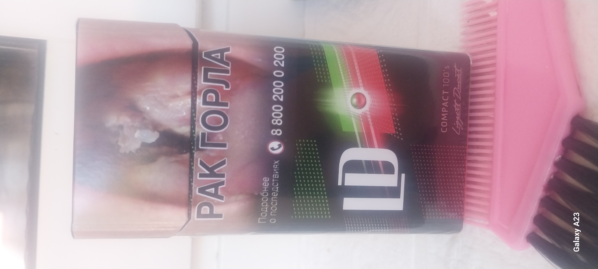 The image displays a cigarette pack, presumably placed on a white surface with a pink hair comb in the foreground. The cigarette pack features prominent text in Russian, including "РАК ГОРЛА" (Throat Cancer) and a phone number "8 800 200 0 200". It also shows the brand logo "LD" and text indicating "COMPACT 100'S Liggett Ducat". The background is indistinct but appears to be an indoor setting. No people are visible in the image. The location in Tuapse, Russia is not directly evident from the visual content.