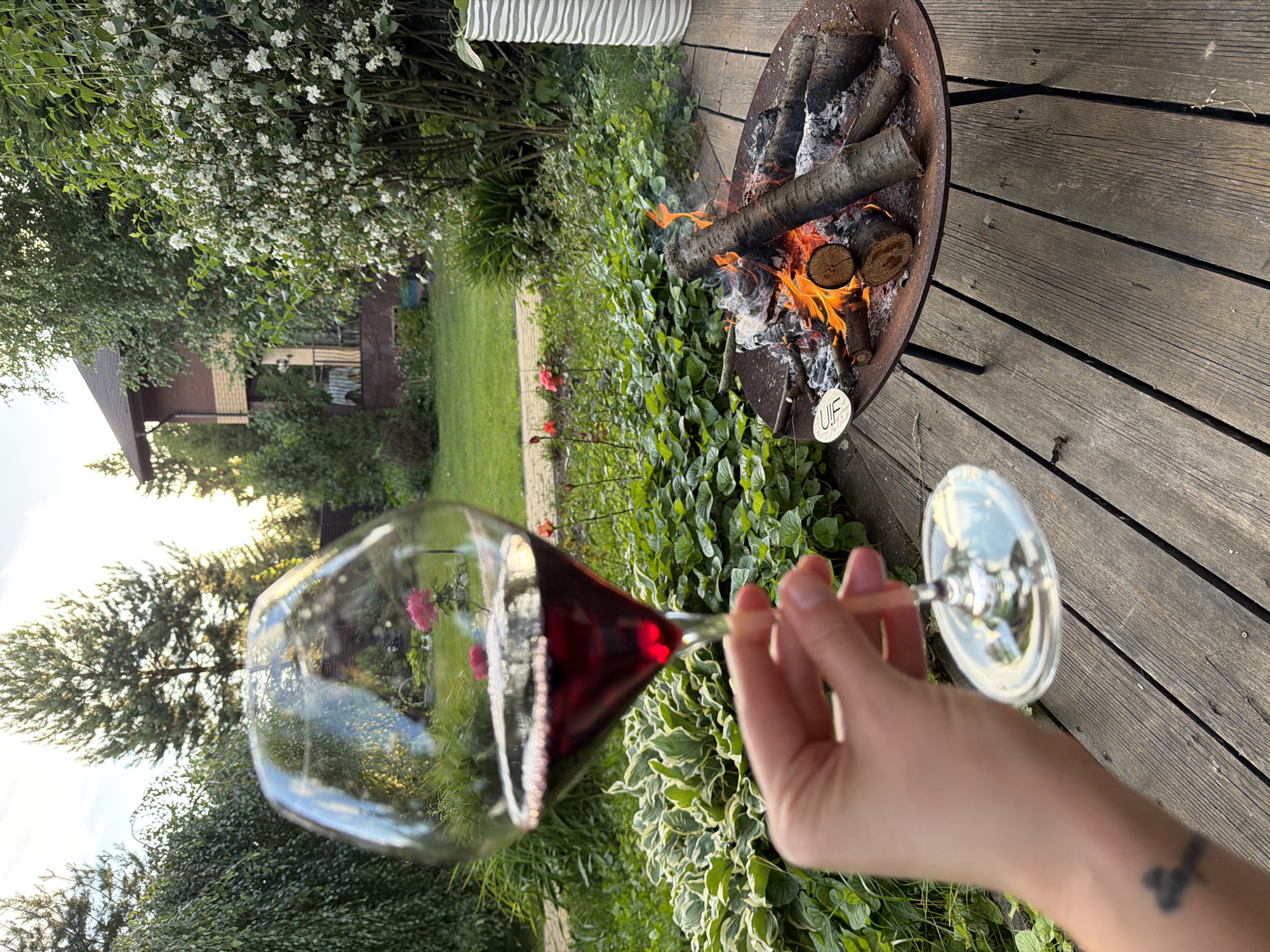The image depicts a backyard scene in Zimyonki, Russia, on a wooden deck with a fire pit. A person's hand is visible, holding a wine glass filled with red wine. The wine glass is in the foreground, reflecting the surrounding environment, including a lush garden with green foliage and flowering plants, as well as a glimpse of a two-story house. In the middle ground, a fire pit contains burning logs and embers, with flames and smoke rising. The background shows trees and the sky, suggesting it is late afternoon or early evening. The overall atmosphere is serene and suggests a moment of relaxation in a domestic outdoor setting.