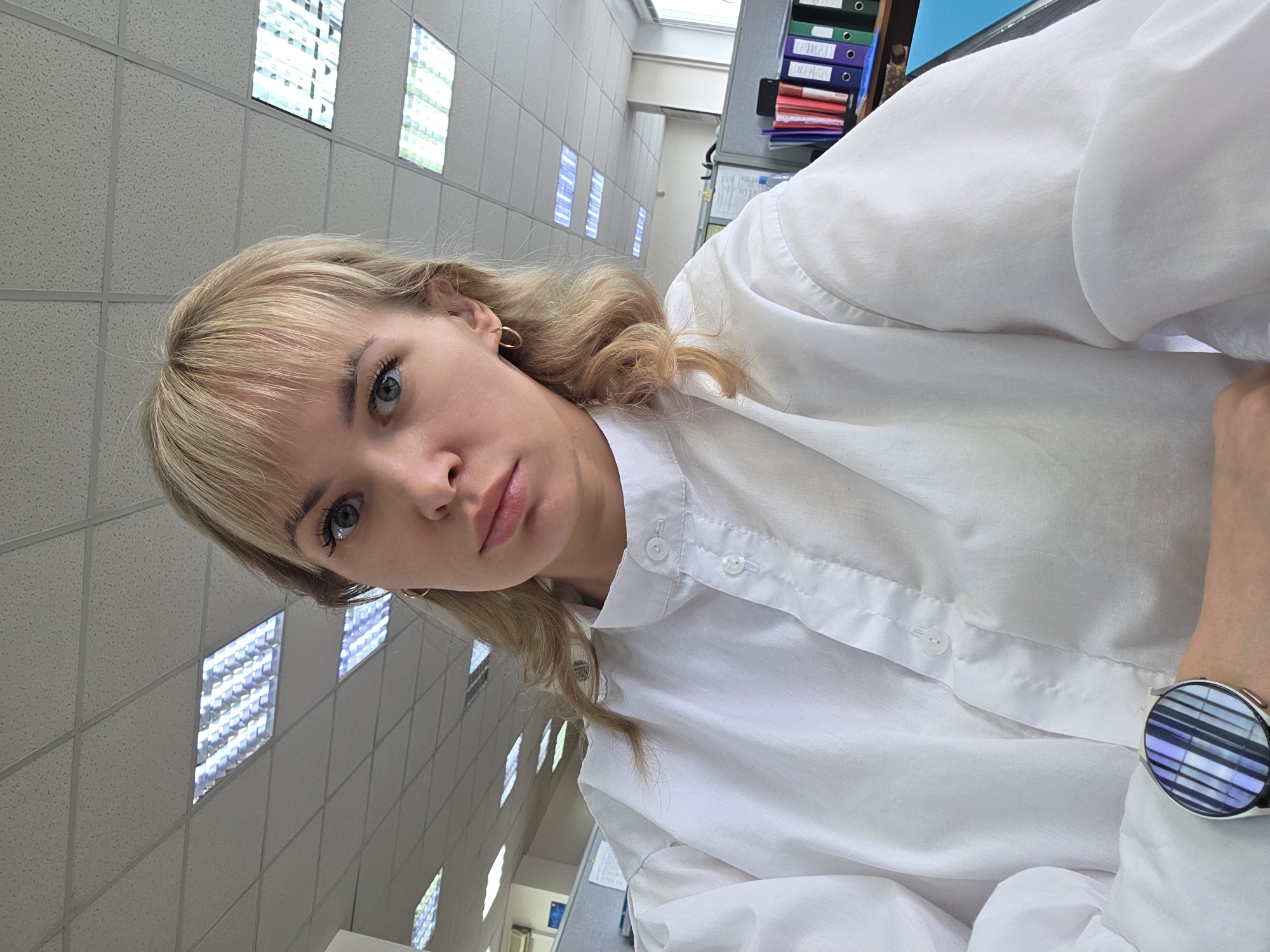 A young woman with blonde hair and blue eyes is the primary subject of the image. She is wearing a white button-down shirt and a watch is visible on her wrist. The background appears to be an office setting, characterized by a grid-like ceiling with recessed lighting fixtures. The presence of filing cabinets with stacked folders in the background further supports the office environment. Based on available data, it's not possible to establish the exact location within Chaikovsky, Russia.