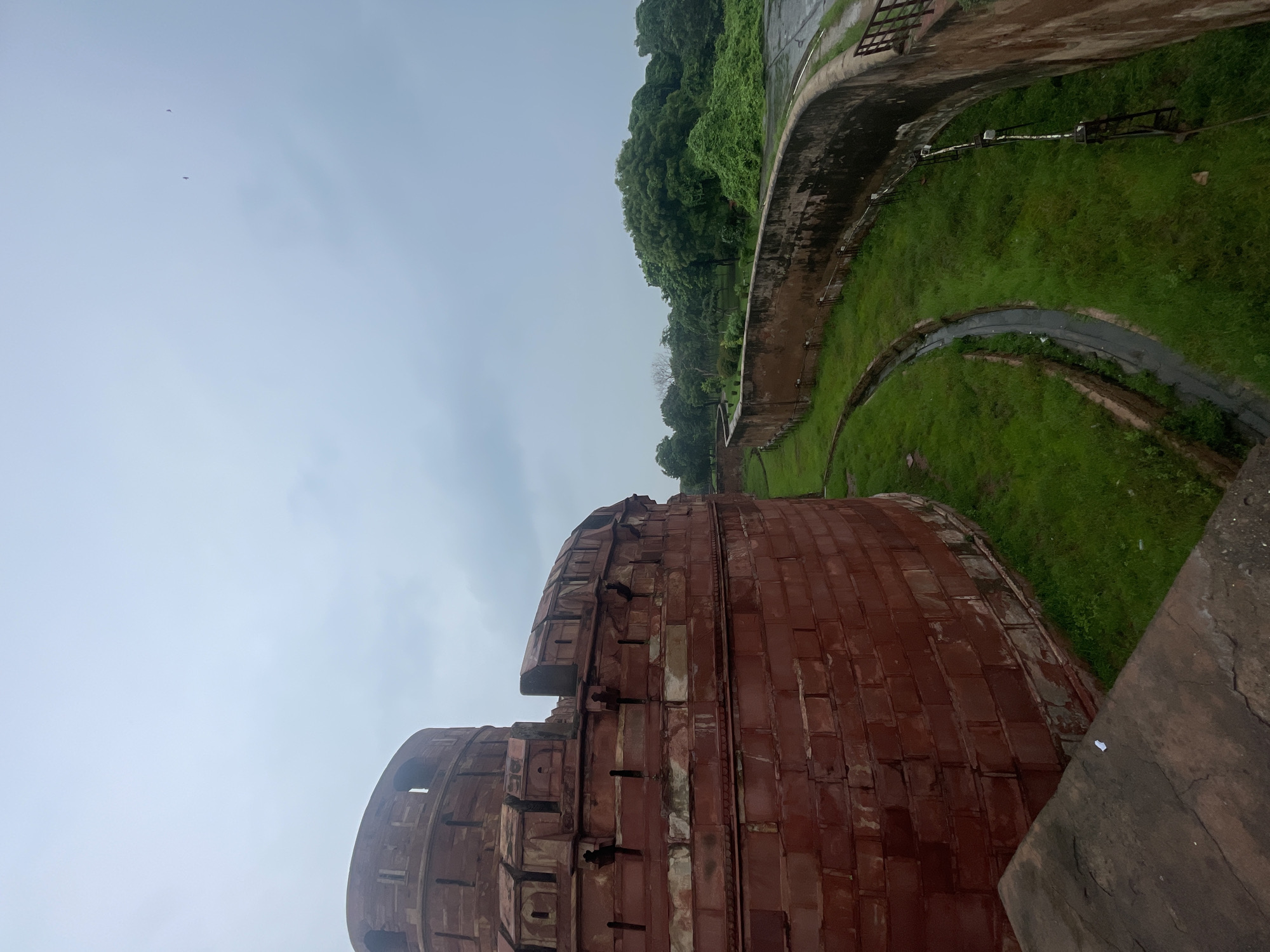The image shows a portion of the Red Fort in Agra, India, on an overcast day. The foreground features a curved, red sandstone wall, characteristic of Mughal architecture, with crenellations and small openings at the top. To the right of this wall, a pathway follows the curvature of a lower, weathered brick wall, beyond which is a lush green grassy area with a narrow channel or drain. In the background, a dense line of trees creates a natural border. The sky is a uniform, light gray, suggesting a cloudy or possibly rainy atmosphere, with a couple of distant birds visible. The overall scene depicts an architectural and natural landscape within the historical context of the Red Fort.