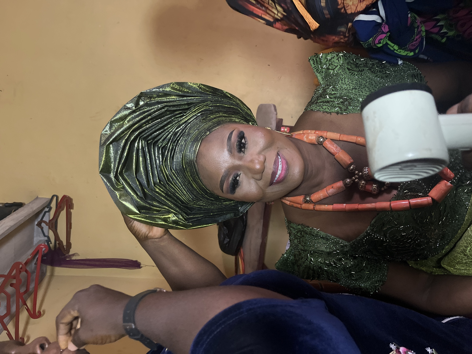 The image features a woman adorned in traditional Nigerian attire, suggesting a celebration or special event. She wears a green, intricately folded headwrap (gele) with a shimmering gold undertone and a matching green dress with beaded details. A coral beaded necklace is prominent around her neck. She is smiling and appears to be in the process of having her makeup applied or adjusted, as a hand holds a white object (possibly a makeup tool) near her face.
In the background, the wall is painted a pale yellow. Other individuals are partially visible, indicating she is likely surrounded by others preparing for the occasion. A clothing rack with hangers is visible on the wall. The image's specific location within Warri, Nigeria, cannot be determined solely from the visual information provided.