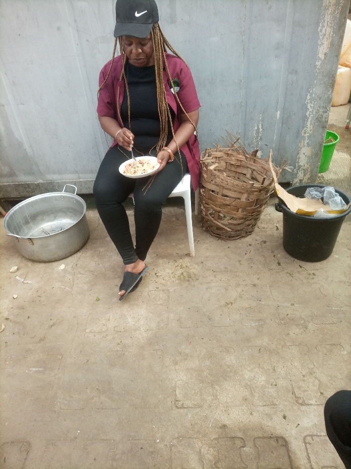 An adult female is seated on a white plastic chair, engaged in eating a meal from a white plate held in her left hand, while using a silver fork with her right. She wears a black Nike baseball cap, a dark reddish-brown open-front shirt over a black top, black leggings, and black open-toed slide sandals. Her hair is styled in long, light brown braids. To her immediate left on the ground is a large, circular, metallic pot. To her right, a large, cylindrical woven basket sits beside a black plastic bucket containing crumpled items. The background features a light grey wall with visible wear, and further right, several translucent yellow plastic containers and a green bucket are visible. The ground is a light-colored, patterned concrete or tiled surface. The scene is located in the city of Opete, Nigeria.