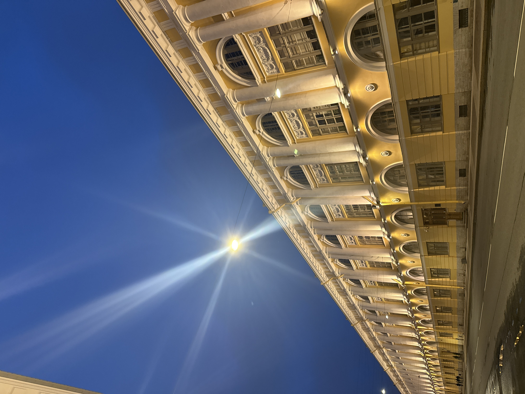 The image depicts a building with classical architecture, featuring a long facade with rows of columns and arched windows, illuminated at night. The building is painted in shades of yellow and cream. A streetlight is visible, creating a bright flare and streaks in the sky. The sky is a deep blue color. A street runs along the side of the building, with reflections visible on the pavement. Based on the architecture, the location is likely in Saint Petersburg, Russia, potentially near the General Staff Building on Palace Square. There are no people visible in the image.