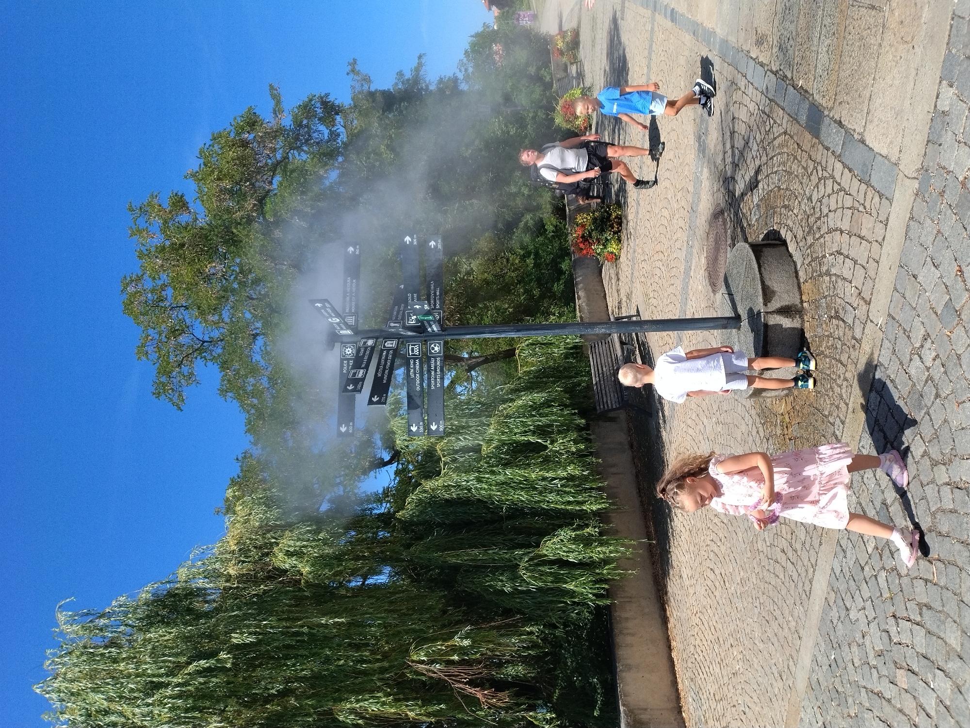 In Písek, Czechia, on a bright sunny day, a misting fan is active, cooling a cobblestone pathway. A young girl in a pink dress walks towards the viewer, holding a purple object. Behind her, a boy in a white shirt and shorts stands facing away. To the right, a man in a grey t-shirt and black shorts, carrying a backpack, walks past. A boy in a blue t-shirt and white shorts walks ahead of him. A weeping willow tree is on the left, with a park bench visible behind it. A directional signpost with multiple arrows and text in Czech is positioned near the misting fan. Lush green foliage and red flowers in planters add to the park-like atmosphere.