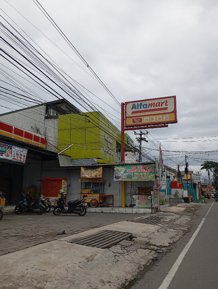 The scene is located at the coordinates -6.994 latitude and 107.555 longitude in Katapang, Indonesia. The image captures a street lined with buildings and shops. A prominent sign for "Alfamart," a convenience store chain, is visible on the left side of the street. The sign is mounted on a pole and is white with red and yellow lettering. The pole extends up into a cloudy sky and is encircled by a dense network of power lines. The buildings are two-story and have a mixture of brick and painted surfaces. The facade of the building behind the sign has a green and yellow paint scheme, while the building to the left is a single-story structure with a red, yellow, and white paint scheme. In front of the building, a group of three motorcycles are parked. They appear to be parked on a concrete slab, which slopes gently down to the street. There are additional shops and businesses along the street, including a "Tahu Goy" stand with a yellow and red sign. A sign for "Es Teller" can be seen on the right side of the street. The street is paved with asphalt and is bordered on each side by a curb and a walkway. The street is mostly empty, with only a few vehicles visible in the distance. The overall scene is one of a bustling commercial district.