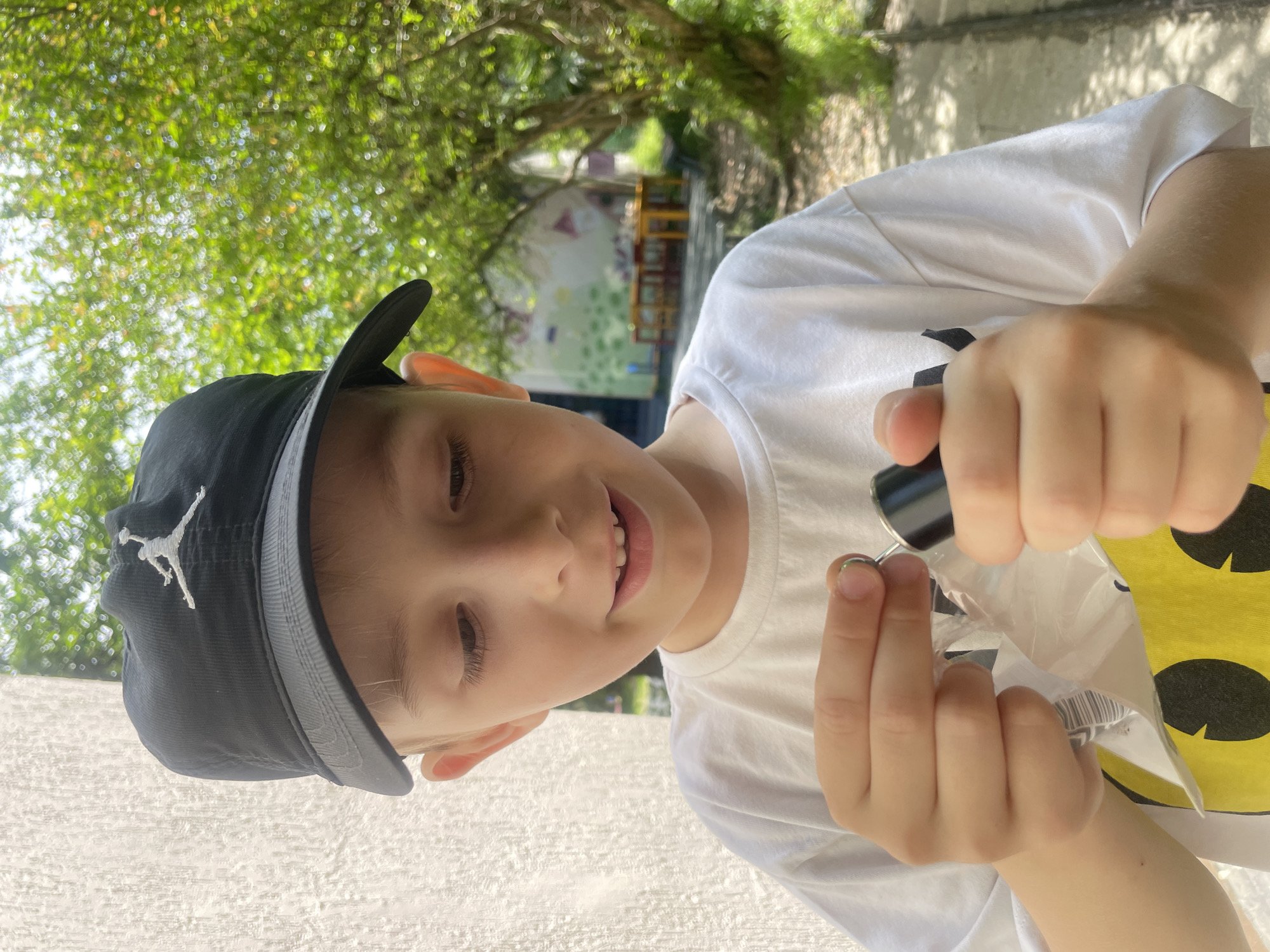A young boy with blond hair, wearing a black baseball cap with a white Jumpman logo and a white t-shirt with a yellow smiley face design, is holding a small, black cylindrical object with a silver tip in his hands. He is looking down at the object with a focused expression and his mouth is slightly open. The background shows a textured white wall on the left, blurred green foliage and trees on the right, and a hint of a colorful mural and outdoor tables in the distance. The lighting suggests it is daytime, with sunlight casting shadows. There is no visible indication of the city of Sochi, Russia in the image.