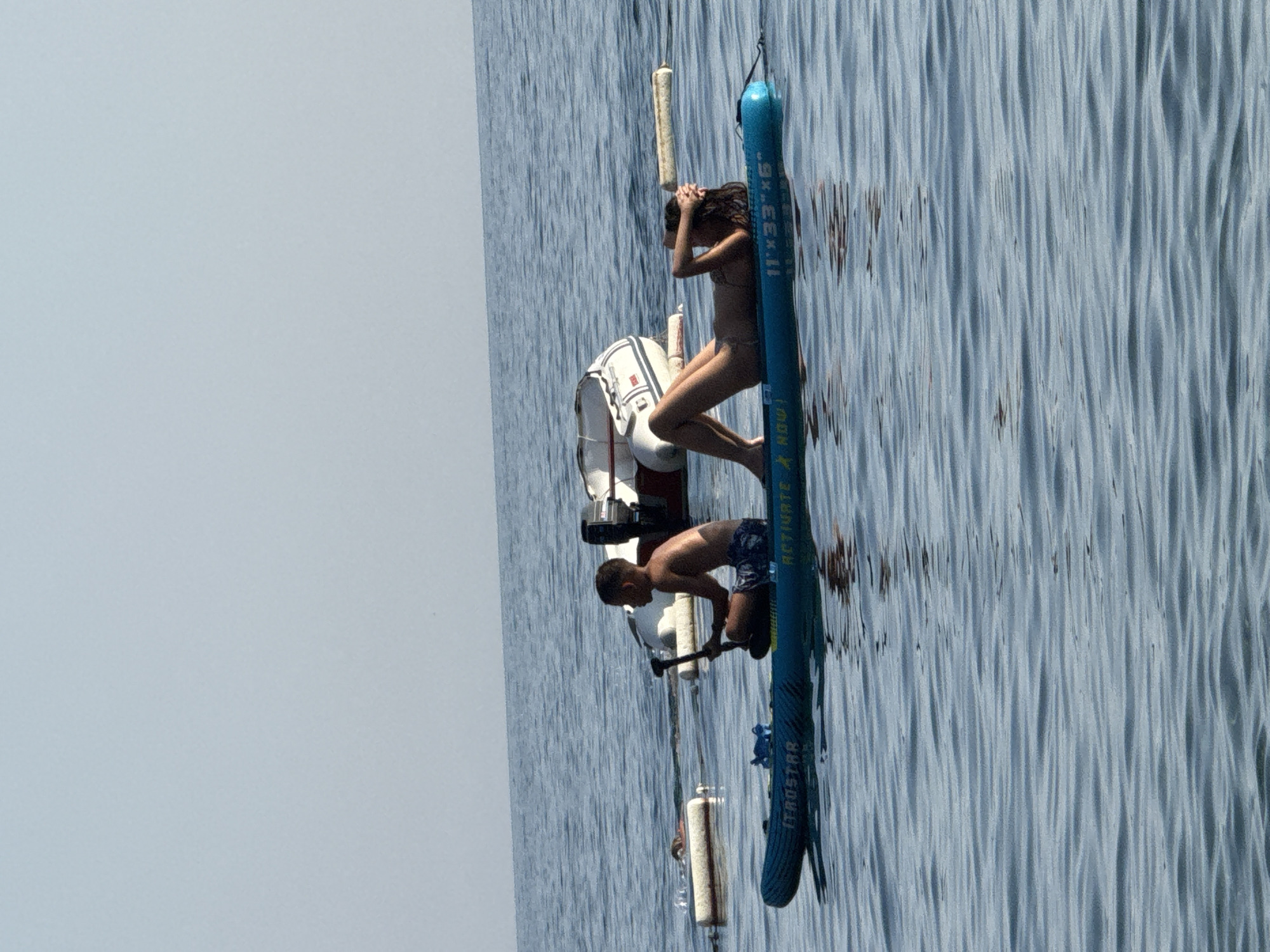 An image captures a serene scene on the Adriatic Sea near Sveti Stefan, Montenegro. Two individuals are positioned on a blue inflatable paddleboard. On the left, a young boy sits cross-legged, holding a paddle. On the right, a woman reclines on her back, her hands behind her head. The paddleboard is labeled with "ITROSTAR" and "ACTIVATE NOW". In the background, a white inflatable boat with an outboard motor is partially visible, along with several white buoys. The water is calm with gentle ripples, reflecting the clear, pale blue sky.
