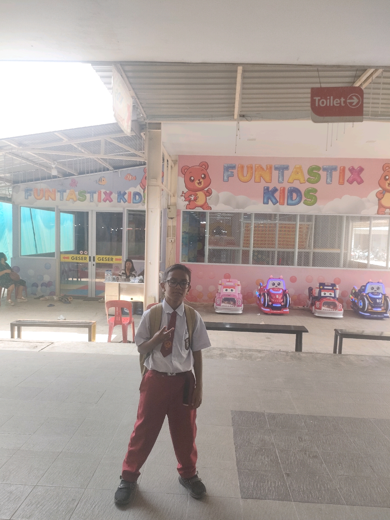 A young boy wearing a school uniform, consisting of a white shirt, red pants, and a red tie, stands posing in front of a building. The building has a sign that reads "FUNTASTIX KIDS" in colorful letters. To the right of the boy, several colorful kiddie rides shaped like cars are visible. A "Toilet" sign with an arrow pointing to the right is mounted on the ceiling. The setting appears to be an outdoor area or a covered walkway of an entertainment venue, possibly in Batam, Indonesia, given the context of "FUNTASTIX KIDS," a known indoor playground chain. The boy is looking at the camera and has his right hand raised in a peace sign. The ground is tiled, and benches are placed in the background.