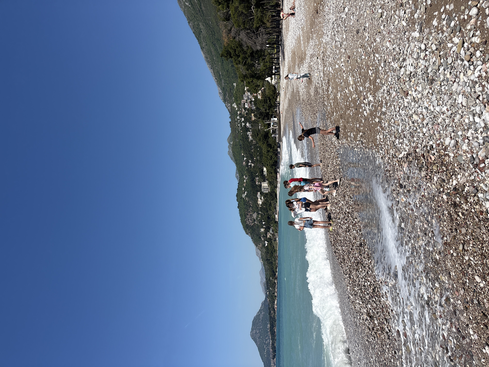 A horizontal outdoor shot presents a beach scene in Bar, Montenegro, under a clear blue sky. The terrain transitions from the sky on the left to a pebble beach on the right. On the far left, the sky is a gradient of blue. Adjacent to the sky, a mountain densely covered with green trees runs vertically along the left side of the image. Houses can be seen nestled within the trees along the mountainside. At the bottom, turquoise water meets the pebble beach, forming white waves.
A group of people are standing at the edge of the water. Further up the beach, a person in a black shirt stands with outstretched arms. In the distance, there's another person further up the beach. The beach itself is composed of pebbles in various shades of brown, grey, and white. The scene is brightly lit by sunlight.