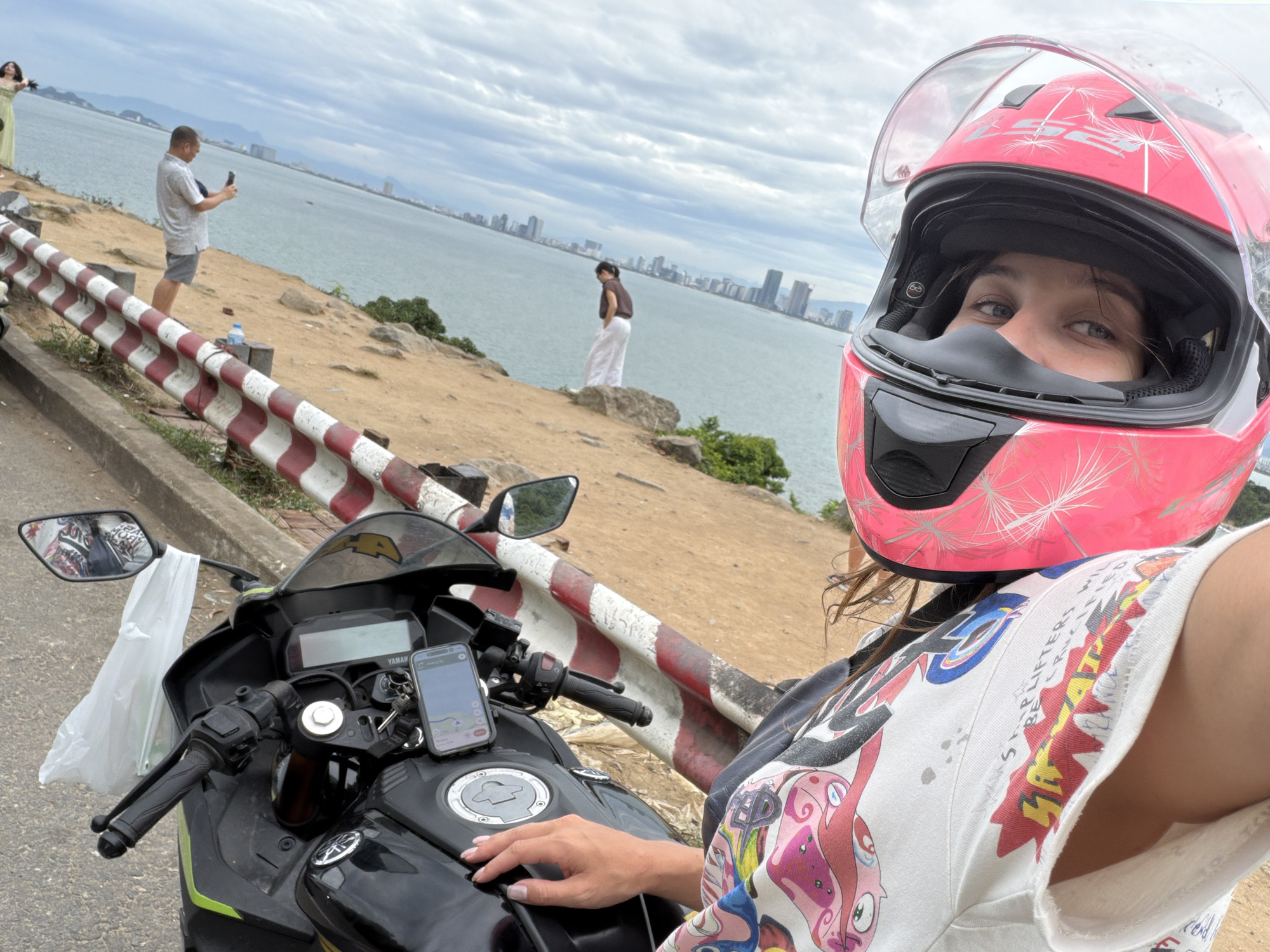 A woman wearing a pink helmet with a clear visor is taking a selfie while sitting on a black motorcycle parked on a roadside overlooking the city of Da Nang, Vietnam. The motorcycle has a phone mounted on the handlebars displaying a navigation app. In the background, the vast sea stretches to the horizon, with a panoramic view of Da Nang's cityscape featuring numerous tall buildings and skyscrapers. Several people are visible in the middle ground; one man is taking a photo of the scenery with his phone, and another person is standing near the edge of the cliff. The roadside is lined with a red and white striped barrier. The sky is overcast, with scattered clouds.