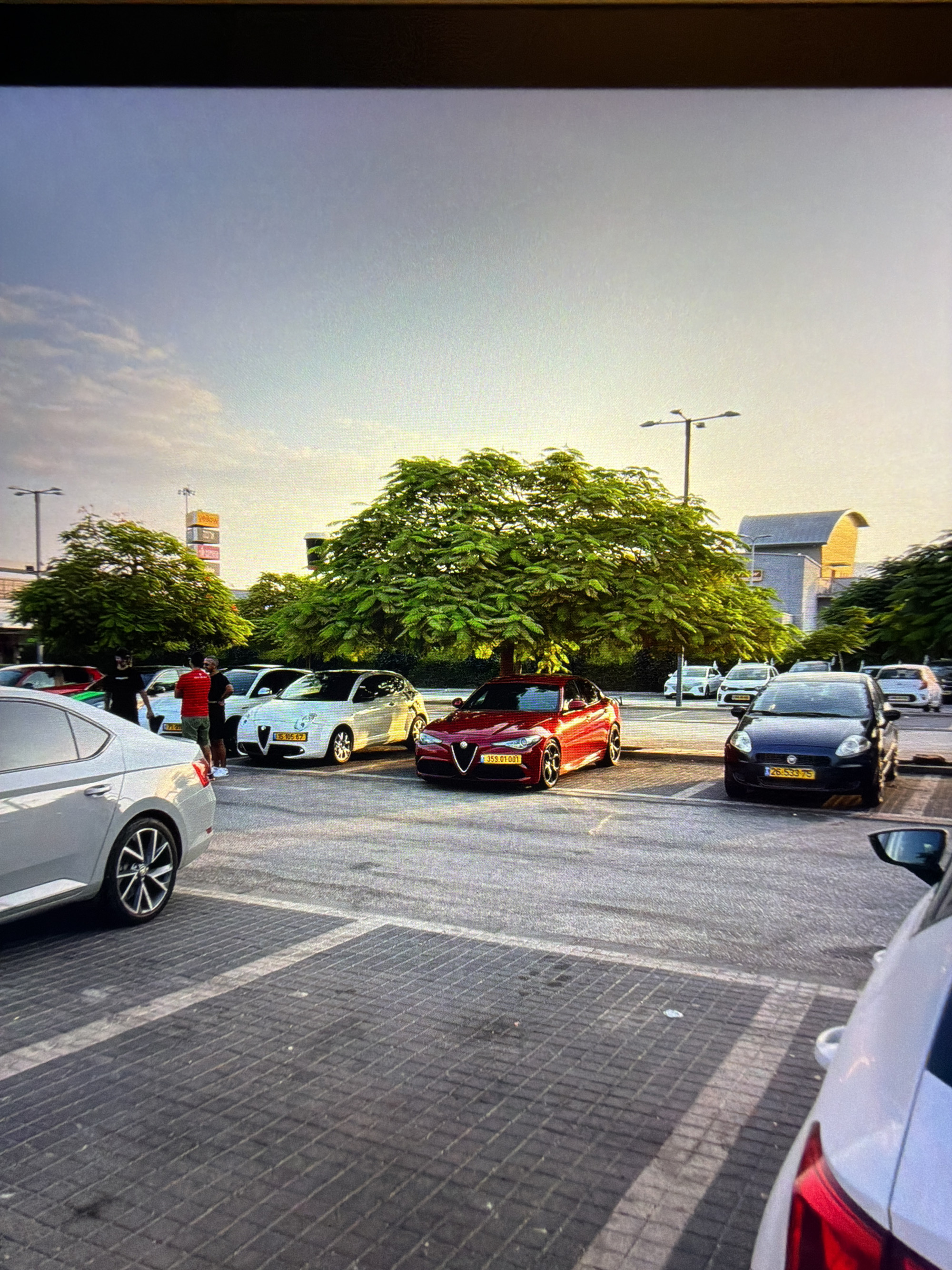 The image depicts a parking lot in Kafr Yasif, Israel, with several vehicles parked and a few individuals present. The lighting suggests it is late afternoon or early evening, with sunlight casting long shadows across the patterned pavement.
In the foreground, a silver sedan is parked, facing away from the camera. To its right, a red Alfa Romeo sedan is parked, with a yellow license plate reading "359 01 001". Behind the red car, a white hatchback with a yellow license plate is visible. Further to the right, a dark blue Fiat hatchback is parked, also with a yellow license plate.
Two men are standing near the white hatchback. One man, wearing a red shirt and khaki shorts, is facing the camera. Another man, wearing a black t-shirt, is standing beside him, facing away. A third person, partially visible, is also in the vicinity.
In the background, more cars are parked, including white sedans and possibly a red vehicle. Several trees with lush green foliage are scattered throughout the scene, providing shade. A lamppost is visible to the left, and a multi-story building with brightly colored signage stands in the distance, suggesting it might be a commercial area or a shopping center. The sky is partly cloudy.