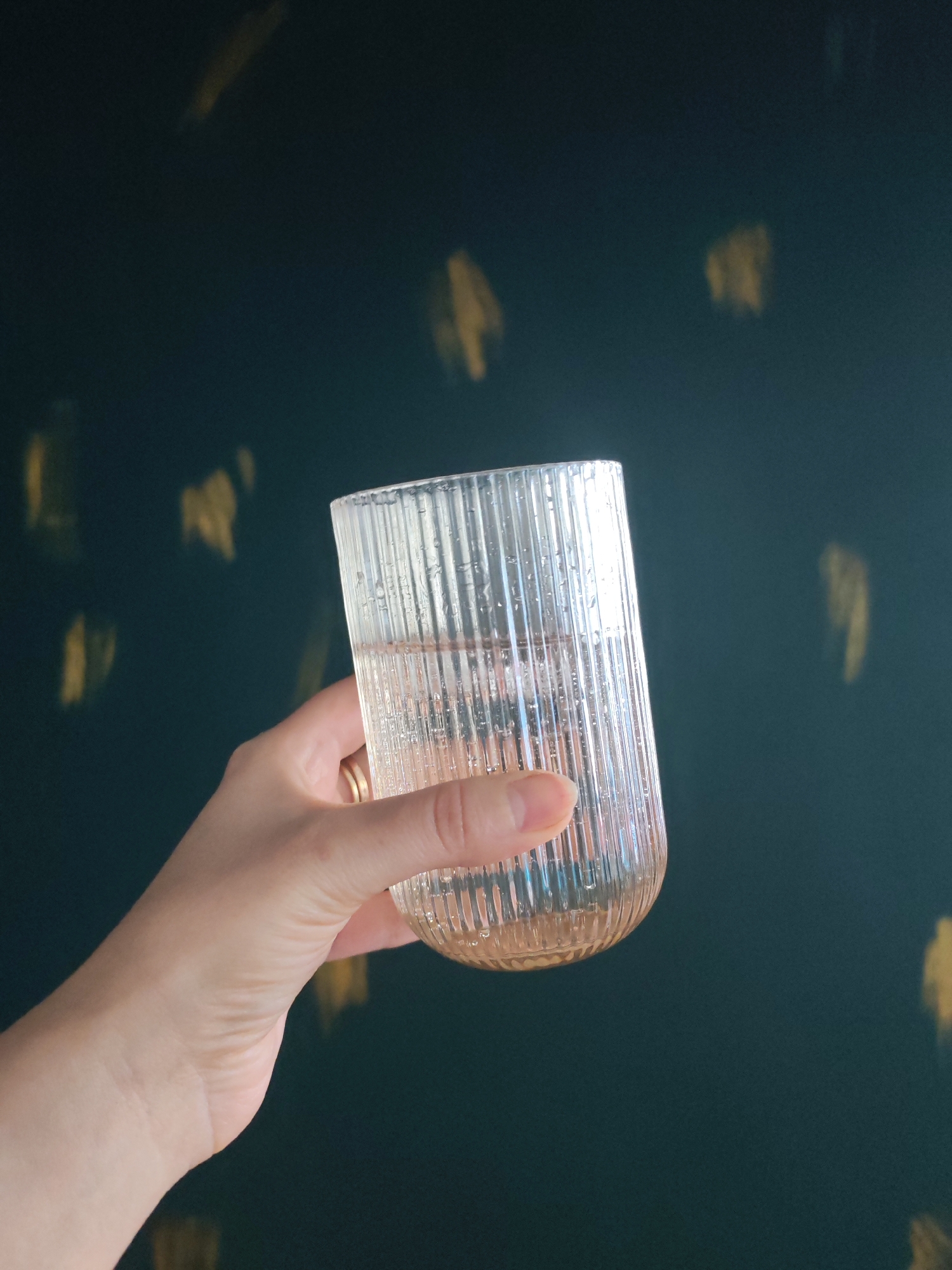 A hand holds a ribbed glass filled with a light-colored liquid against a dark teal background with blurred yellow splotches. The glass is positioned in the center-right of the frame, and the hand is visible from the wrist down, with a ring on the ring finger. The background appears to be a wall with some sort of decorative paint or lighting effect. There are water droplets on the outside of the glass. The image is a medium shot, focusing on the hand and the glass. The overall impression is one of simple elegance and a focus on the drink and the vessel.