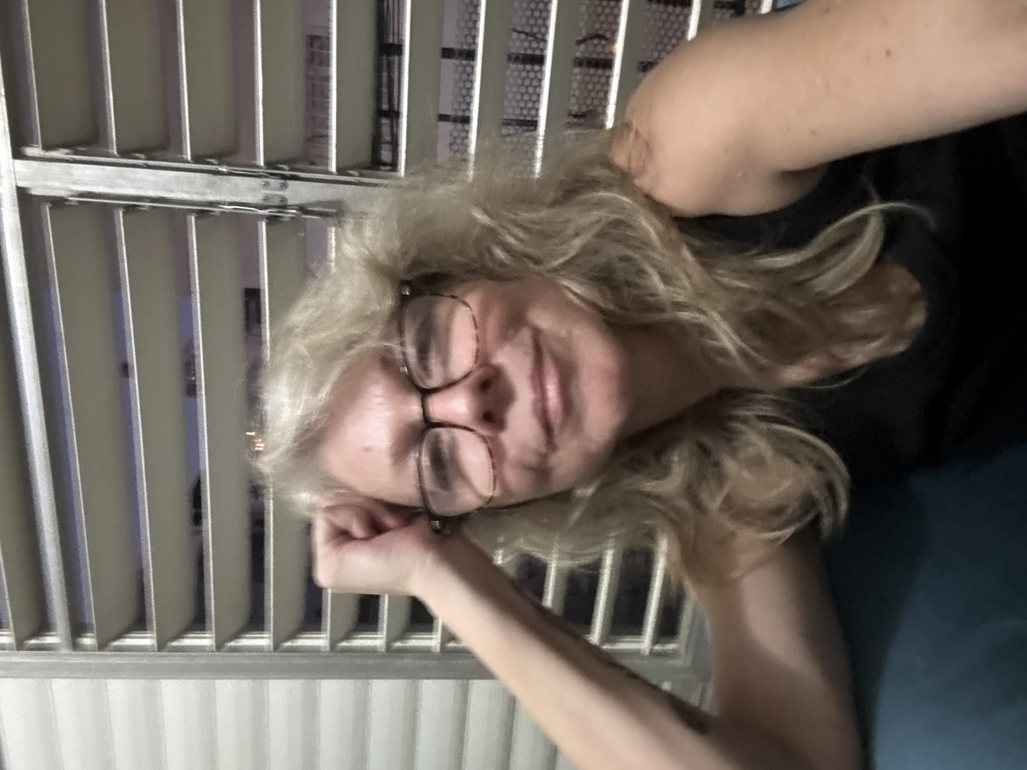 An older Caucasian woman with blonde hair and glasses is pictured indoors, leaning her head on her hand. She is wearing a black tank top and is seated on a blue couch. Behind her is a window with louvers that are partially open, revealing a dark interior. The scene is dimly lit, suggesting it is either evening or nighttime. The woman's pose and facial expression suggest a relaxed or contemplative mood. The location is identified as Ramat Gan, Israel, though no specific landmarks or contextual clues are visible in the image to confirm this.