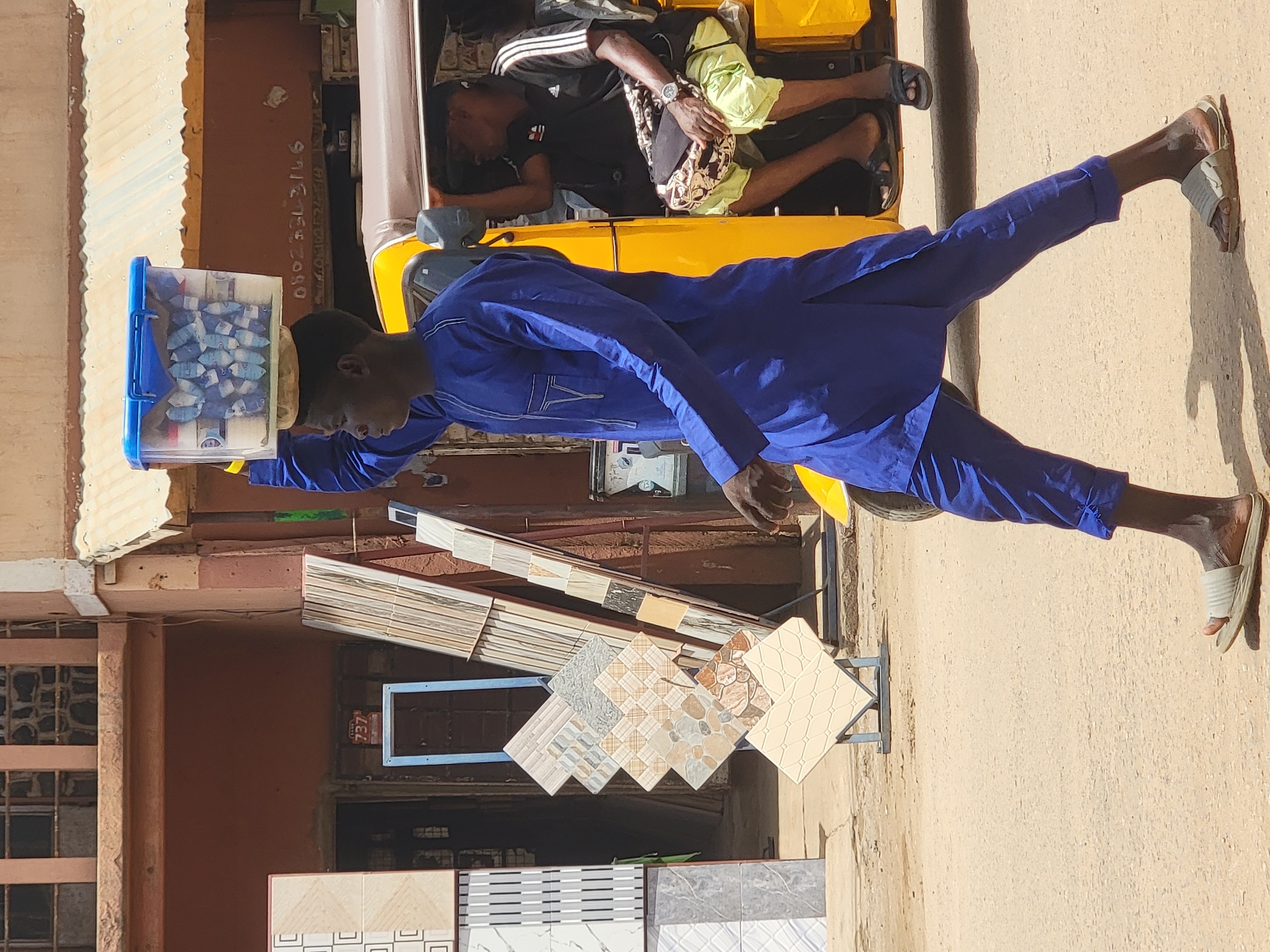 A street scene in Maiburiji, Nigeria, featuring a man in a blue outfit balancing a blue-lidded container on his head. Another person lies in a yellow motorized tricycle behind him. Ceramic tiles are displayed on a rack near a building with bars on the windows.