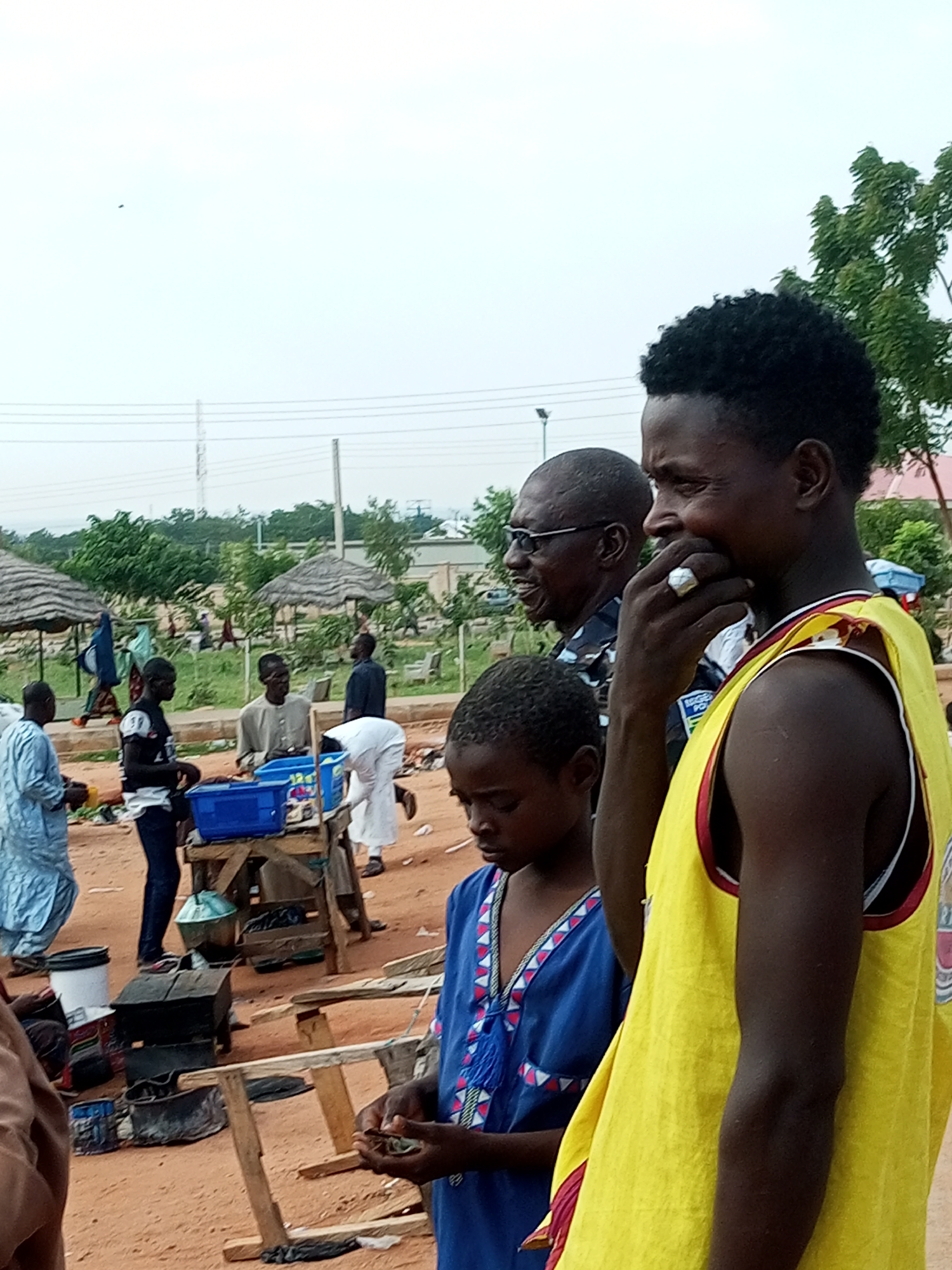 The image depicts an outdoor scene, likely in the city of Gombe, Nigeria, characterized by a dusty, sandy ground and scattered vegetation. Several individuals are present, some engaged in transactions or discussions. In the foreground, a young boy wearing a blue garment with red and white trim is holding something in his hands, looking down. To his right, a man in a bright yellow sleeveless top with red trim is shown in profile, with his hand near his mouth and wearing a ring. Behind them, a man with glasses and a dark shirt, possibly a police officer due to a patch on his sleeve, is looking to his left. Further in the background, other people are visible, including a man in a light-colored robe, a woman in a blue headscarf and dress, and individuals near thatched-roof stalls. Various objects are scattered around, such as blue plastic containers, wooden tables and benches, buckets, and what appears to be cooking equipment. The overall atmosphere suggests a market or a community gathering in an open area.