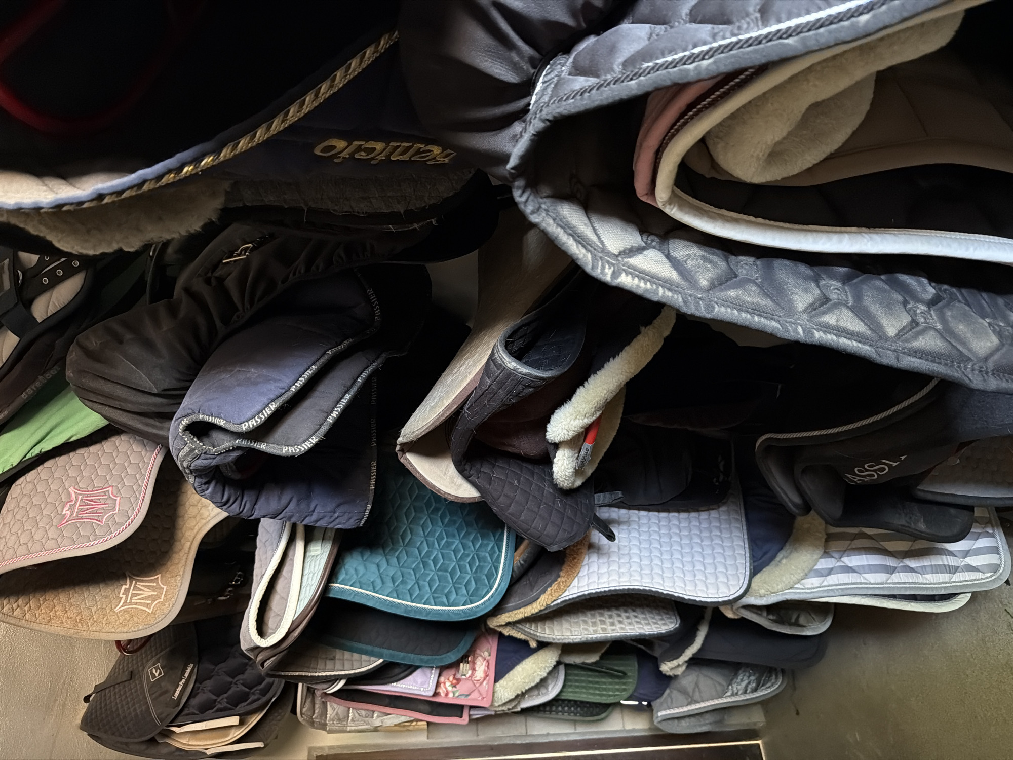 The image depicts a dense collection of horse saddles and related equipment stored in what appears to be a stable or tack room. The saddles are layered on top of each other, showcasing a variety of colors, textures, and patterns. Some saddles are plain black, while others feature decorative stitching, quilted designs, or colored trim. There are also visible saddle pads with various patterns, including one with a floral design and another with a textured hexagonal pattern. The overall impression is one of organized storage, with the equipment neatly arranged for accessibility. The lighting suggests an indoor environment, possibly a barn or a dedicated tack room. There are no people visible in the image.