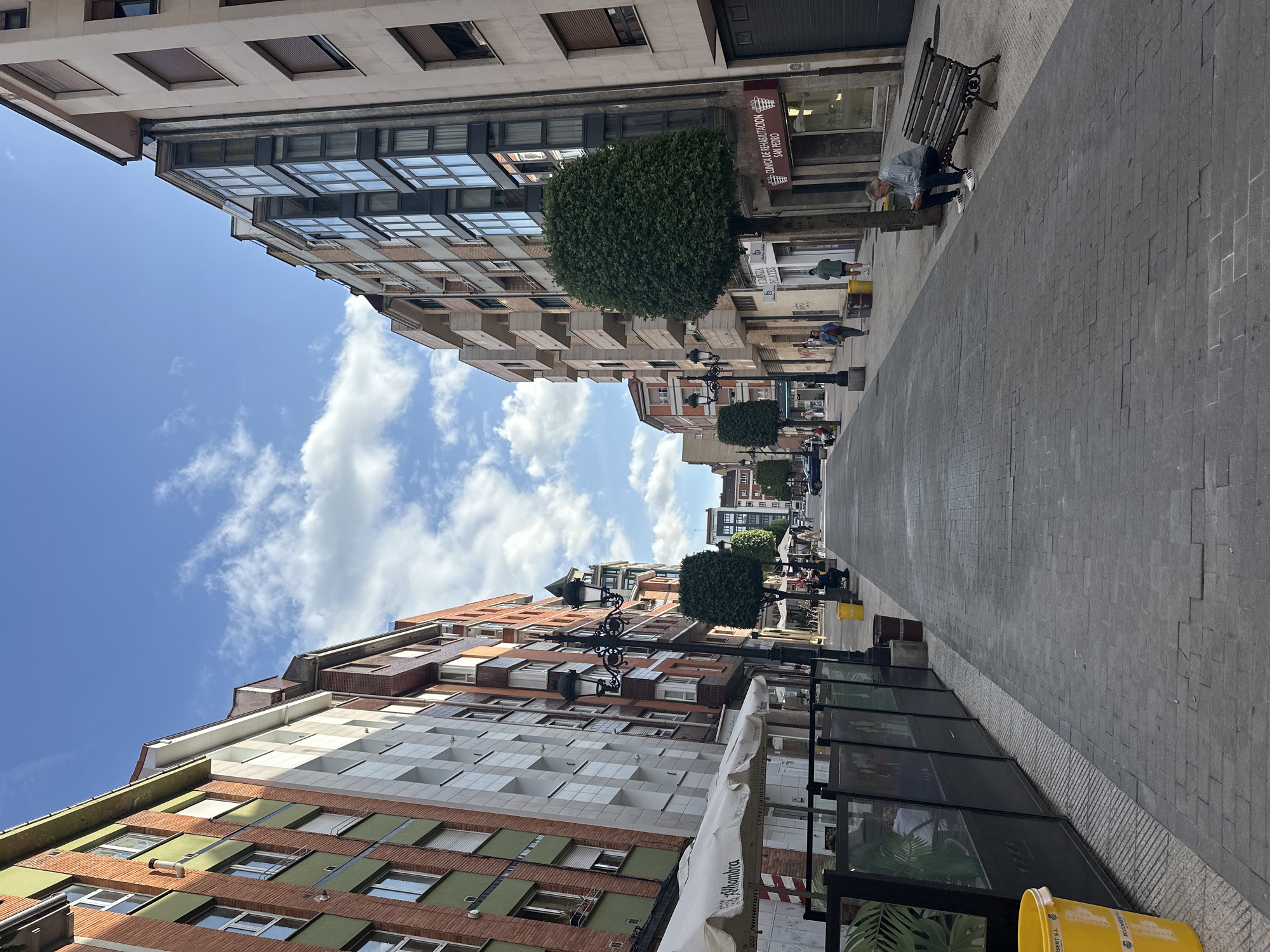 This image captures a vibrant, sunny street scene in Oviedo, Spain, looking down a wide pedestrian pathway lined with multi-story buildings. The sky is bright blue with scattered white clouds, indicating pleasant weather, likely during the mid-day or afternoon.
On the right side of the street, several buildings display diverse architectural styles. The closest building features a light-colored facade with dark-framed windows and internal blinds. Below it, a large, rounded green bush or small tree is planted. Adjacent to this, a modern building boasts a prominent glass facade with square window panes. Further down, another building is characterized by a series of lighter-toned, staggered rectangular balconies. A red sign on one of these buildings clearly displays the text "CLÍNICA DE REHABILITACIÓN SAN PEDRO" (San Pedro Rehabilitation Clinic). A person is seen sitting on a dark wooden bench with ornate metal legs on the sidewalk, seemingly engrossed in something in their hands. Other indistinct figures are visible walking further down the street.
The left side of the street also features various buildings. One structure has a distinctive facade with green panels and brown brick sections, while another displays a pattern of grey and white squares. A decorative black streetlight with multiple lamps stands prominently on this side. There appears to be an outdoor seating area, possibly belonging to a cafe or restaurant, indicated by a partially visible white awning and glass barriers along the sidewalk. A yellow bucket, possibly for refuse, is visible in the foreground with "ALHAMBRA" printed on its side, a common Spanish brand.
The street itself is paved with grey block tiles, typical of pedestrian zones. The overall atmosphere is that of a calm and active urban environment.