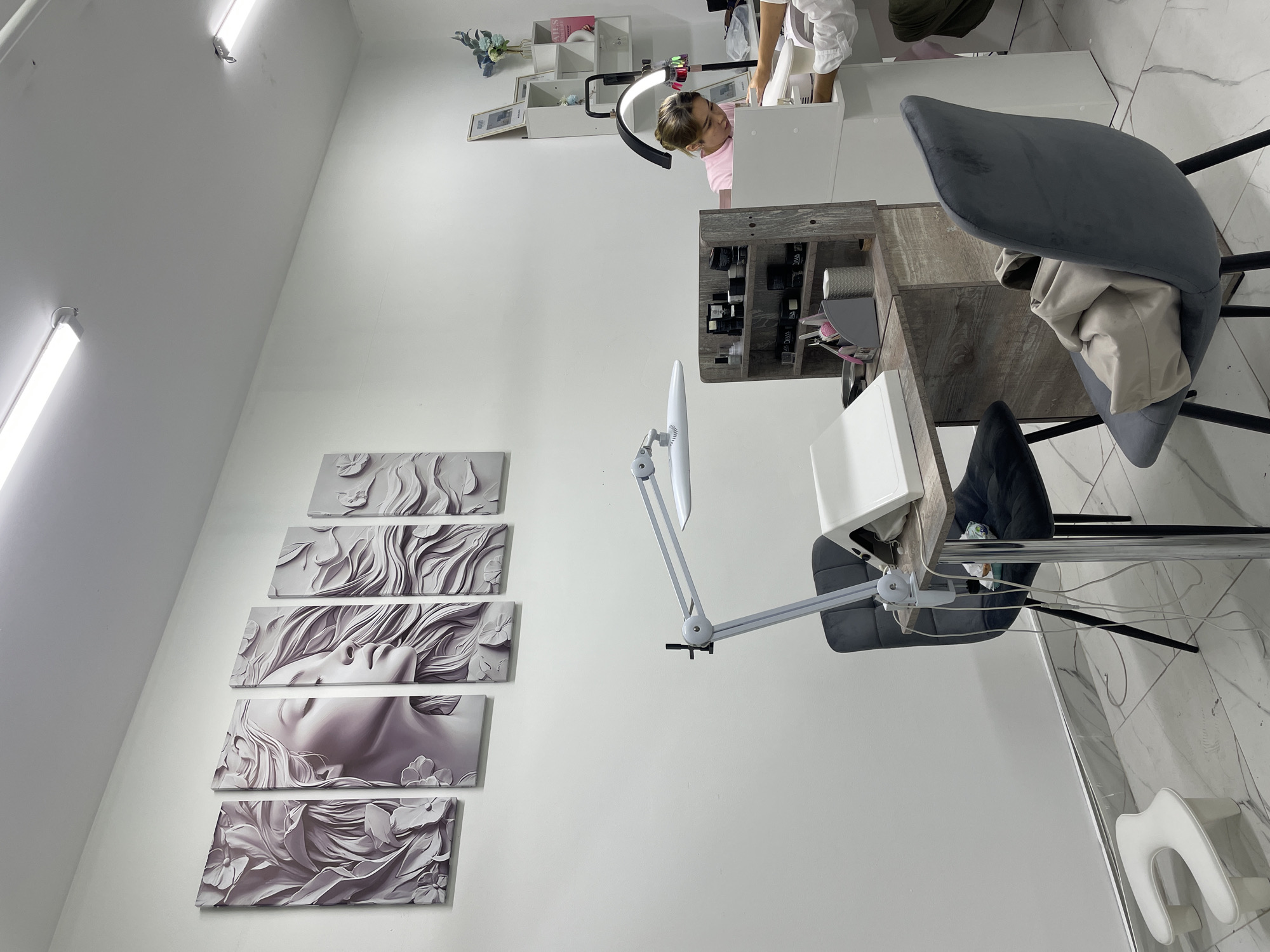 This image depicts the interior of a nail salon, likely in Almaty, Kazakhstan, as indicated by the presence of a "Diva Nails" product on a shelf, a brand known to operate in the region. The scene is well-lit with overhead fluorescent lights. Dominating the left wall is a five-panel artwork featuring a stylized woman's face and floral elements in shades of gray and purple.
In the foreground, a black upholstered chair with chrome legs is positioned at a nail desk. On the desk, a white nail lamp with a flexible arm is clamped to the side, along with a white client rest and a gray organizer containing various nail tools and products, including bottles labeled "Diva." A smaller white footrest is situated on the tiled floor to the left.
To the right, another nail desk area is visible. A woman wearing a pink top is seated and appears to be working on a client's nails, with her hands visible. A tall, multi-tiered shelving unit behind her holds various nail polish bottles, decorative items, and framed pictures. The walls are painted white, and the floor is covered in marble-patterned tiles. A gray upholstered chair with a light-colored bag draped over it is positioned near this second workstation.