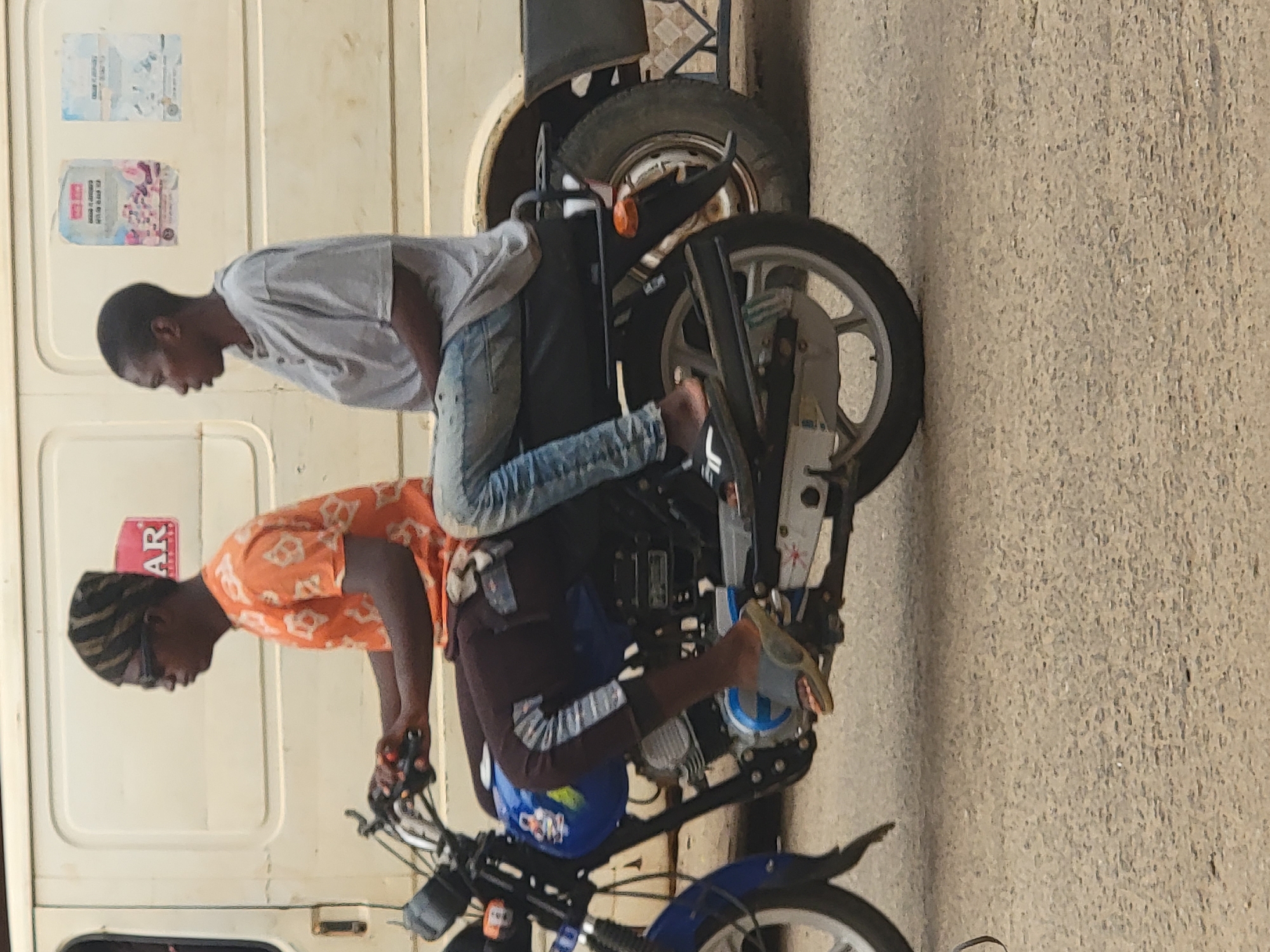 Two men are riding a motorcycle in what appears to be Maiburiji, Nigeria. One man in an orange shirt is driving the motorcycle. The passenger behind him is wearing a gray shirt and blue jeans. The motorcycle is blue and black. A white van is next to them. The road is made of asphalt.