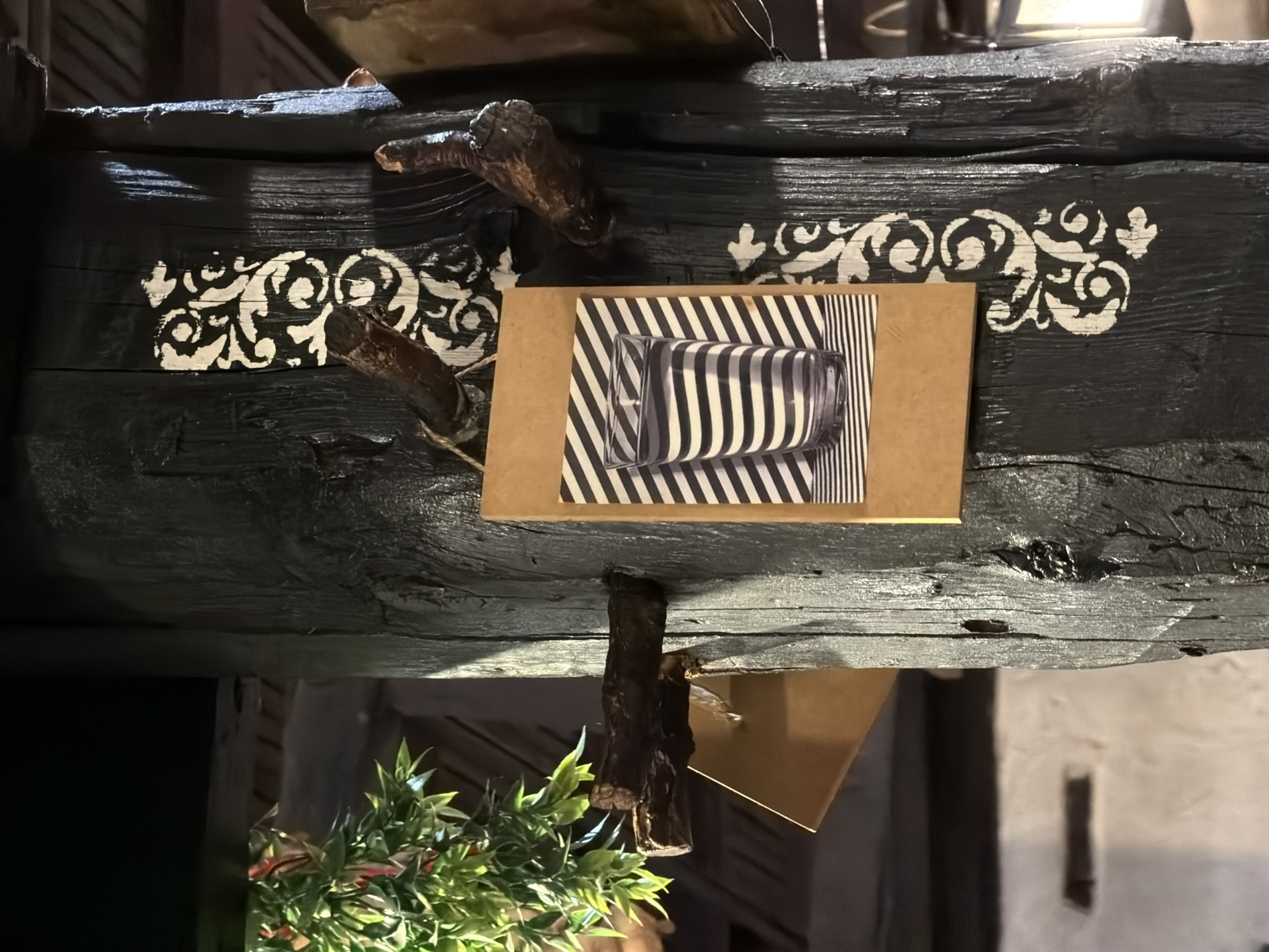 An interior scene in Varna, Bulgaria, is dominated by a robust, vertically oriented wooden pillar, stained a dark, almost black color, exhibiting visible grain and textured surface irregularities. Several gnarled, dark brown branches protrude horizontally from the pillar at various points. White decorative stencil patterns, featuring intricate scrollwork and ornate motifs, are applied directly onto the dark wood in segmented vertical strips. Centrally affixed to the pillar is a rectangular piece of light brown cardstock, which frames an image. This image displays a clear, cylindrical glass tumbler with horizontal black and white stripes, resting on a surface patterned with diagonal black and white stripes. To the upper left, a cluster of artificial foliage with vibrant green and hints of red leaves is visible. The background reveals dark wooden structural elements in the upper section and a segment of a white wall with a dark, rectangular recess or opening in the lower left. A partial view of another object, brown in color, and a light source are discernible on the far right.