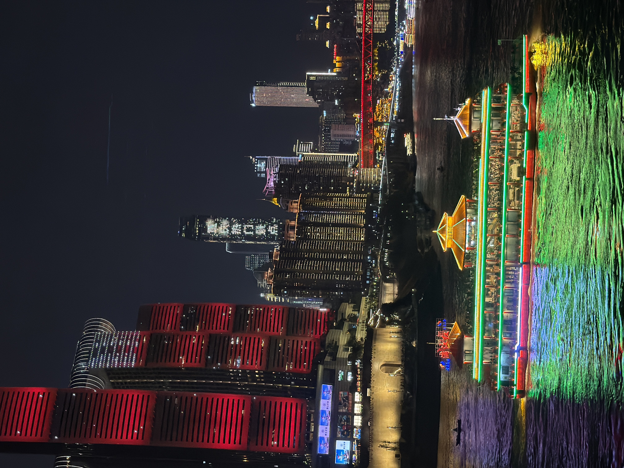 This nocturnal image captures a section of the cityscape in Nan'an District, China, viewed from an elevated perspective overlooking a wide river. In the immediate mid-ground, a large, multi-decked leisure boat, adorned with prominent green and red LED lighting along its hull and decks, navigates the dark water. The boat features traditional Chinese-style pagoda roofs, illuminated in golden-yellow, on its upper structures. Numerous individuals are visible on its open decks. To the left of this vessel, a smaller, dark rowboat or similar craft with a single occupant is present on the water.
The background consists of a dense urban environment dominated by illuminated skyscrapers and buildings. On the left, a cluster of modern high-rises stands out, with two exceptionally tall buildings brightly illuminated in solid red, featuring horizontal banding. One building further back displays a distinct white light pattern on its facade, resembling a starburst, and is topped with curved, illuminated structures. Along the riverbank, a fortified stone wall with an arched gateway is visible, illuminated by warm yellow lights, above which further buildings and structures are situated.
To the right of the central buildings, a complex array of skyscrapers and commercial structures glows with various white, yellow, and blue lights. A prominent red-lit bridge spans the river further in the background, connecting different parts of the city. The river's surface reflects the vivid lights from the boat and surrounding structures, creating patterns of green and red. The sky above is dark, with a faint, horizontal light streak visible in the upper right quadrant.