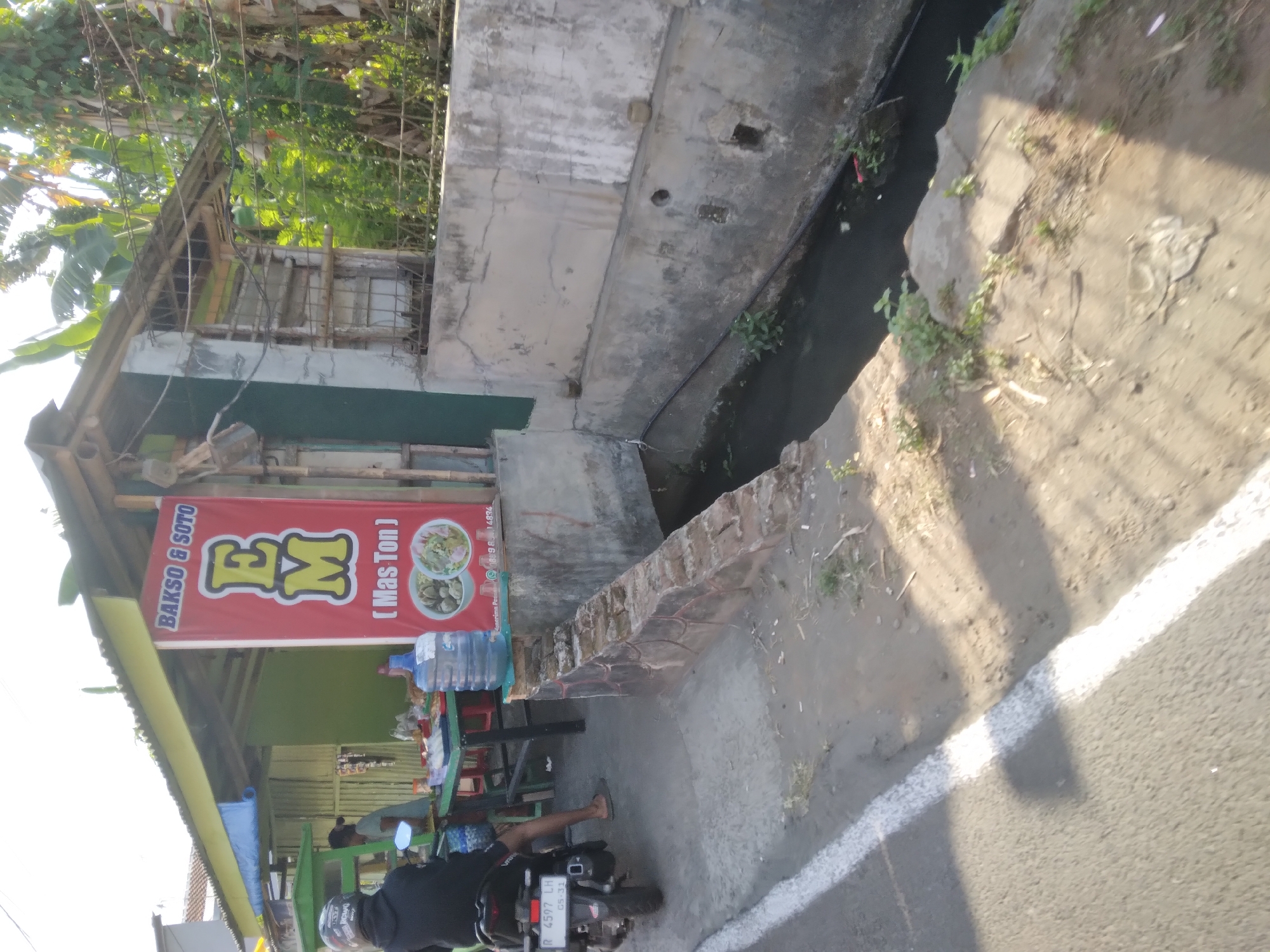 The image shows a street-side eatery in Purwokerto Kidul, Indonesia. A person is seated on a motorcycle parked on the side of the road, facing a stall. The stall has a red banner with the text "BAKSO & SOTO EM (Mas Ton)" and images of food. Behind the stall, there's a structure made of corrugated metal and wood. A large water dispenser is placed on a low concrete wall. The road has a white line marking. To the right of the stall, a dirty drainage channel runs alongside a concrete wall. Vegetation is visible behind the stall and along the drainage channel. The sun is casting shadows, indicating it is likely daytime.