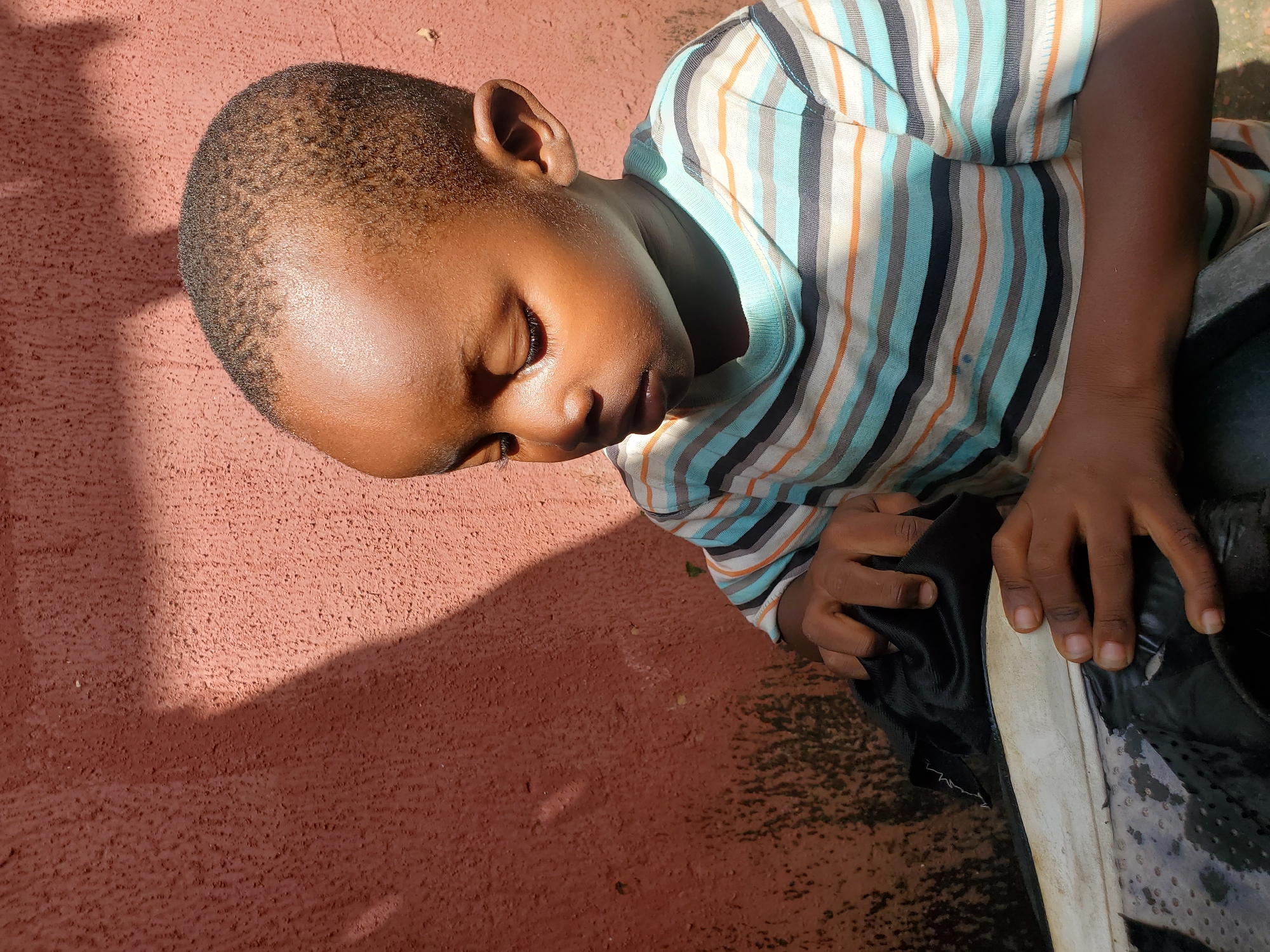 A young boy with dark skin and short hair is the subject of this image. He is wearing a horizontally striped t-shirt with bands of white, light blue, orange, and black. His eyes are closed, and his head is bowed, suggesting he is resting or perhaps feeling tired. His left hand is holding a piece of black fabric, possibly part of his clothing or an object he is interacting with. His right hand rests on a black shoe with a white sole, which appears to be slightly wet or stained.
The boy is positioned on a textured, reddish-pink surface that resembles pavement or a wall. The lighting indicates it is daytime, with shadows cast by an unseen object to the left of the frame, suggesting sunlight. The overall setting appears to be outdoors, possibly in a casual or informal environment in Jos, Nigeria. The boy's posture and closed eyes convey a sense of peace or somnolence. There is no visible text in the image.