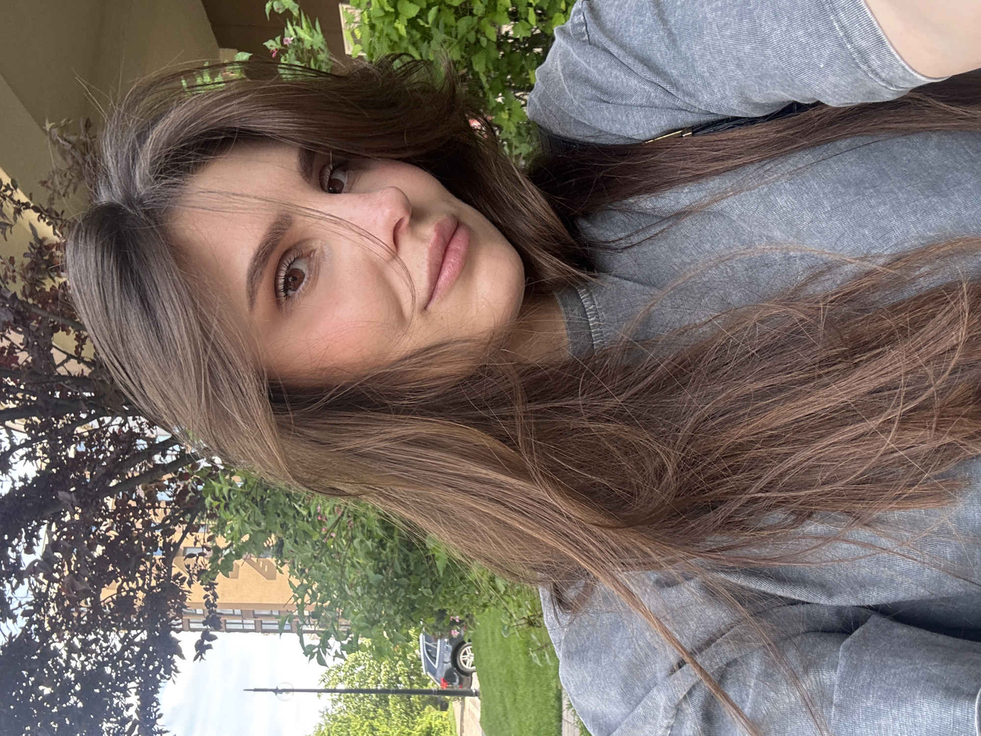 The image features a young woman posing for a photograph in an outdoor setting, likely a residential area in Sofiivska Borshchahivka, Ukraine, during daylight hours. She is the primary subject, angled towards the camera, with long, dark brown hair that cascades over her shoulders and partially across her face. Her brown eyes gaze directly at the camera, and her expression is calm and neutral, with a slight, subtle hint of a smile. She is wearing a simple grey t-shirt.
The background consists of vibrant natural greenery, including dense green bushes and trees with both dark and lighter foliage. Behind the greenery and on the left side of the frame, a light-colored building with visible windows can be seen. Further in the background, a silver car, possibly an SUV or hatchback, is parked on a paved surface adjacent to a grassy lawn. The lighting indicates a clear and bright day. There is no visible text in the image.