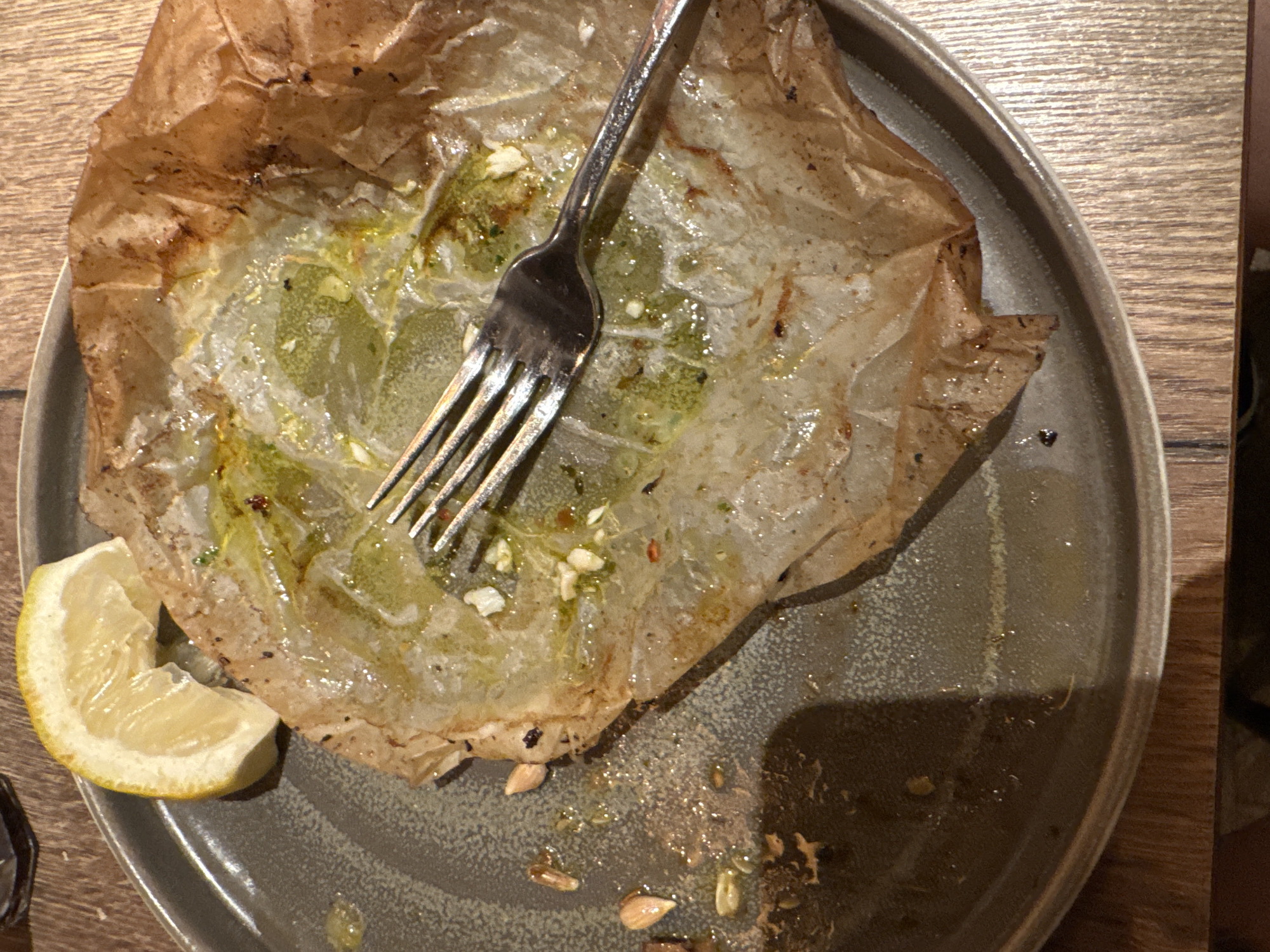 The image shows a close-up, top-down view of a dirty plate on a wooden table. A single fork rests on top of a crumpled piece of parchment paper, which is stained with oil and food remnants. A wedge of lemon is positioned on the left side of the plate. The plate itself is a dark, speckled gray. The lighting suggests an indoor environment. The image does not provide any information about the city of Varna, Bulgaria.