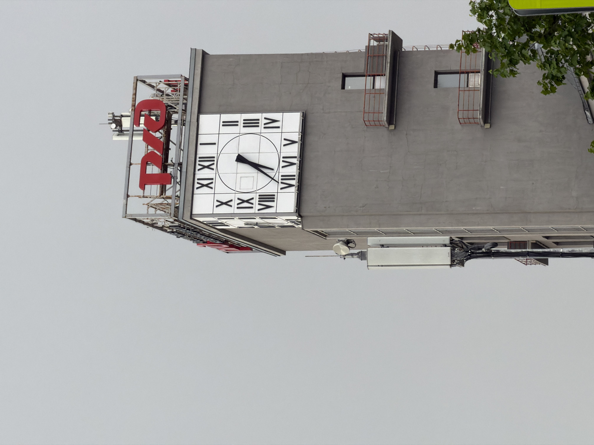The image shows the headquarters of Russia Today (RT) in Moscow, Russia. The gray building features the RT logo in red lettering on the side and a large clock face. The clock reads approximately 7:08. The sky is overcast. A tree is visible in the top right corner. Structural elements like balconies and antennae are also visible on the building.