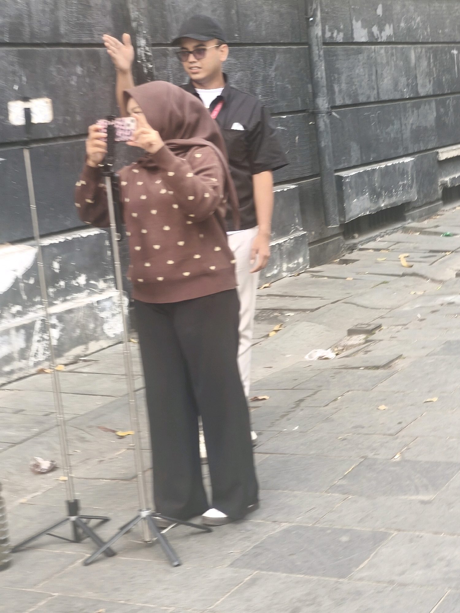 In West Jakarta, Indonesia, two individuals stand on a paved outdoor area, likely a sidewalk or plaza, adjacent to a textured gray wall. A woman in the foreground is holding a pink and black smartphone mounted on a tripod, appearing to record or take a picture. She is wearing a brown hijab and a brown sweater with small yellow polka dots, paired with black pants. To her right and slightly behind her, a man wearing a black cap, sunglasses, a black collared shirt with a white handkerchief in the pocket, and white pants is visible. He has his right arm raised, palm outward, in what could be a gesture of direction or acknowledgement. The ground is paved with square gray tiles, with scattered leaves and debris. Two thin, metallic tripods stand in front of the woman, one with the phone attached.