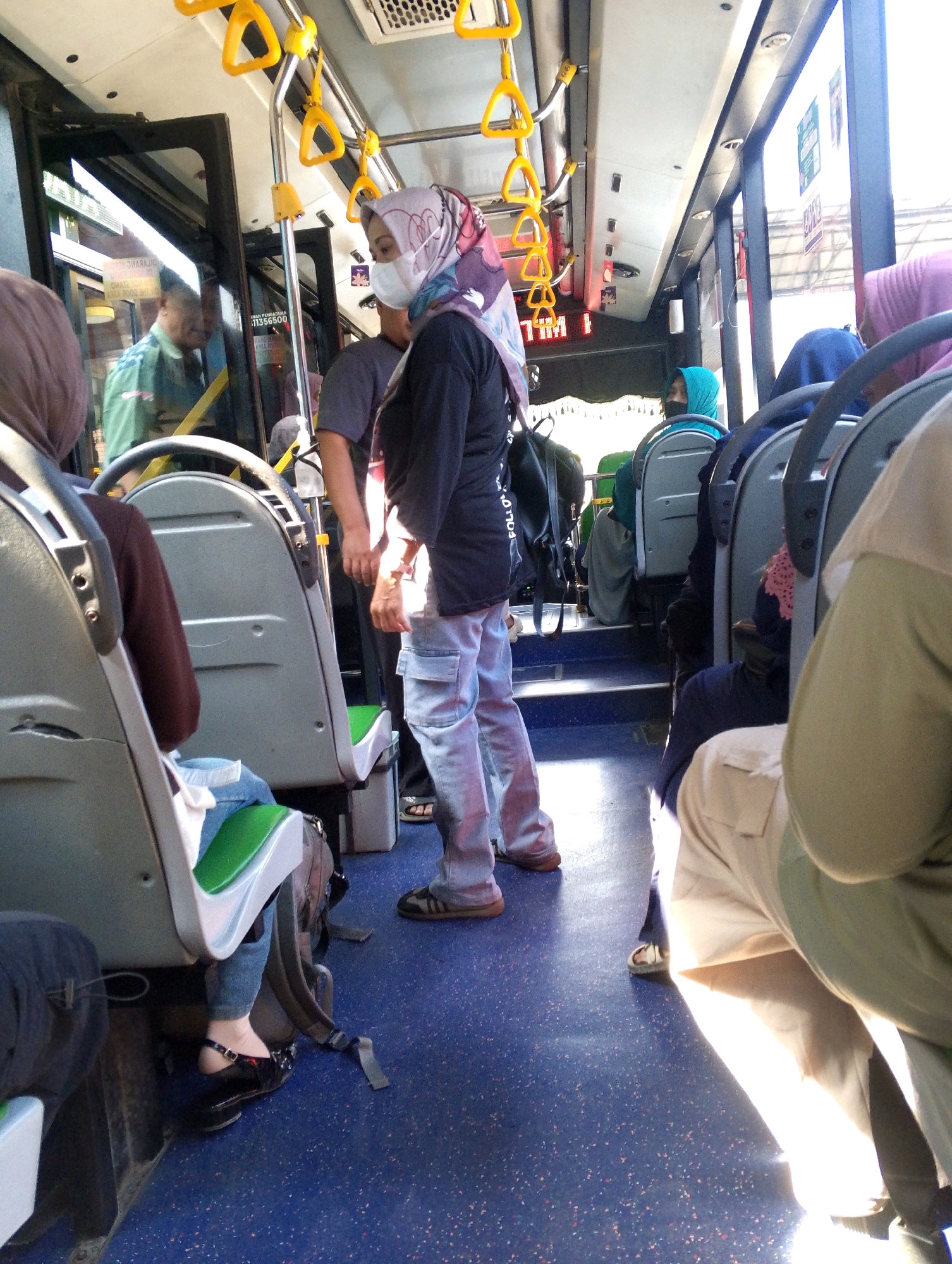 The image depicts the interior of a bus, likely in a city in Indonesia, possibly Meri. Several passengers are visible, most wearing hijabs. One woman in the foreground is wearing a face mask, a black long-sleeved shirt, and light-colored cargo pants. Another passenger in the foreground, to the left, is partially visible and appears to be wearing a dark-colored top and jeans. A man in a green shirt is seen through a glass door, possibly the driver or another passenger. Yellow handrails are installed along the ceiling of the bus. The seats appear to be made of grey plastic with green upholstery. Sunlight streams in through the windows, casting shadows on the blue speckled floor. The overall scene suggests a typical day of public transportation.