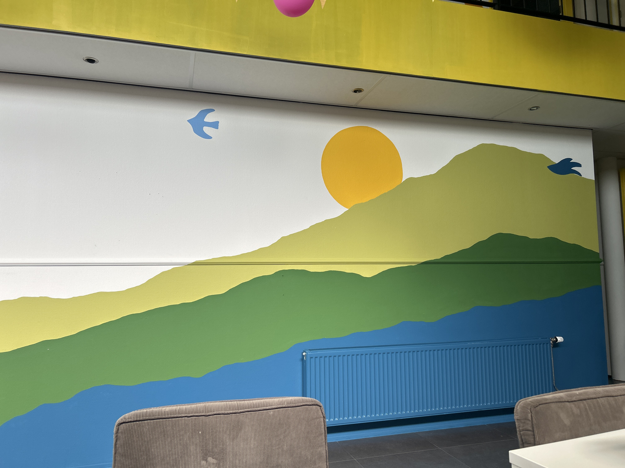 The image shows a mural on a white wall depicting a stylized landscape with rolling hills painted in shades of green and yellow, a blue body of water at the bottom, and a yellow sun. Two small blue birds are painted on the wall, one on the left and one on the right. A bright yellow band runs along the top of the wall. A blue radiator is mounted on the wall in front of the mural. Two upholstered chairs are partially visible in the foreground, and a white table is on the right edge of the image. The location is the reception area of a building in Lisse, Netherlands.