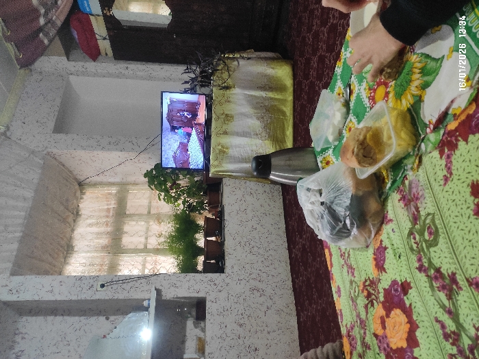 This image depicts an indoor domestic scene in Gulshan, Uzbekistan, recorded on January 16, 2026, at 13:34. The room features textured, light-colored walls and a red patterned carpet. A central rectangular window, fitted with sheer patterned curtains, allows ambient light into the space; multiple potted plants are arranged along its sill. To the immediate left of the window, a wall niche with a shelf contains a luminous object. To the right of the window, a flat-screen television rests on a small, cloth-draped table, displaying an interior scene with a bed and two figures. A dark wooden wardrobe with a reflective surface is visible on the far right. In the foreground, a low table covered with a green and yellow floral tablecloth holds a silver thermos, a clear plastic bag containing unidentifiable food items, a single bun or bread roll, a clear rectangular container with yellow food items, and a smaller clear container with white cubic food items. A person's hands, partially visible on the right, are reaching towards a white dish on the table.