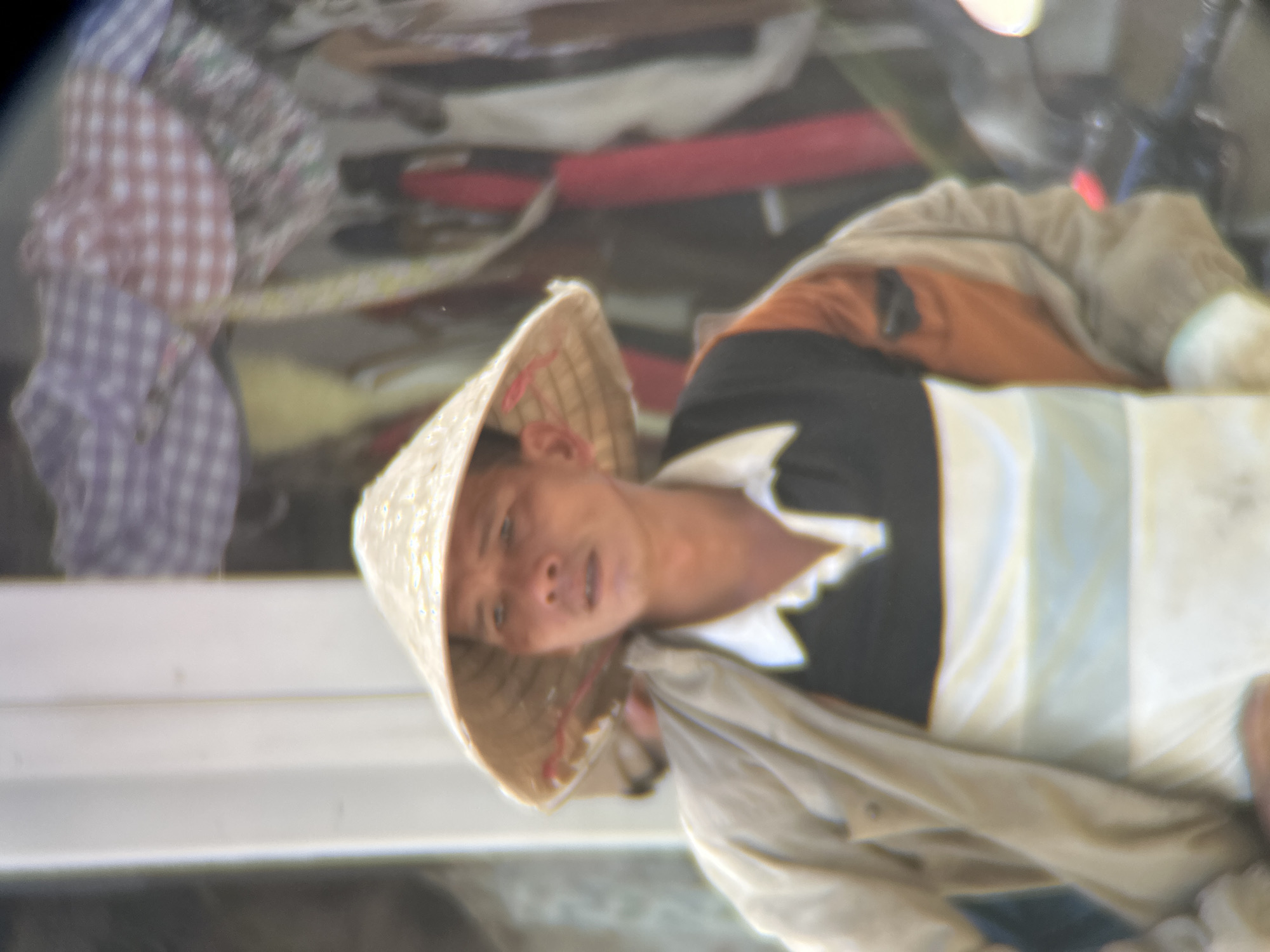 A man wearing a conical hat and a light-colored jacket over a collared shirt stands in front of a blurred background of what appears to be market stalls. The hat casts a shadow over his face, obscuring most of his features. He is looking slightly off to the right of the frame. The clothing of the man is layered and appears to be casual. The background displays a collection of colorful fabrics, possibly merchandise for sale, with some vertical elements that could be racks or poles. The overall lighting suggests an outdoor setting, likely during daytime. The scene is consistent with a marketplace in Đà Nẵng, Vietnam, where such hats and vibrant textiles are commonly found.