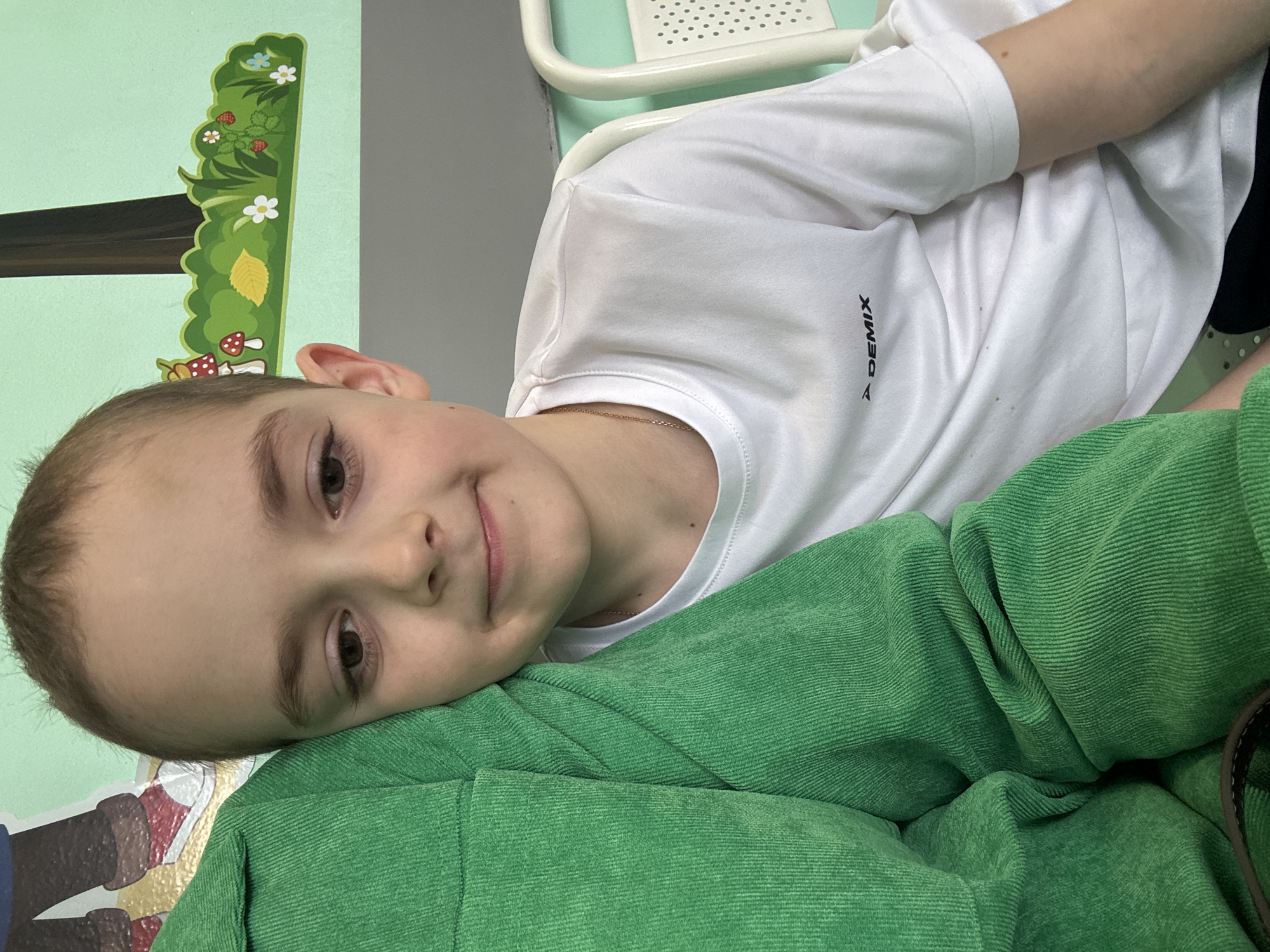 An indoor, eye-level portrait captures a young child, likely a boy, in Klin, Russia. The child, with light brown hair and dark brown eyes, gazes directly at the camera with a gentle, subtle smile. His head is resting comfortably on a vibrant green, ribbed fabric, possibly a cushion or garment.
He is wearing a white short-sleeved t-shirt that displays a black, vertically aligned text logo "DEMIX" on the right side of the chest. A thin gold chain is visible around his neck. The background consists of a light green wall decorated with a colorful, nature-themed border. This decor features lush green foliage, red strawberries, white and blue flowers, a yellow leaf, and red and white mushrooms, suggesting a child-friendly environment such as a playroom or a healthcare setting.
Partial views of other objects are visible: a light-colored, possibly metallic, furniture piece with a perforated pattern in the upper right, and a fragment of another wall decoration depicting a dark shoe with red laces below the child's head. The lighting is bright and even, indicating an indoor daytime setting. The child's calm expression and comfortable pose convey a sense of contentment.