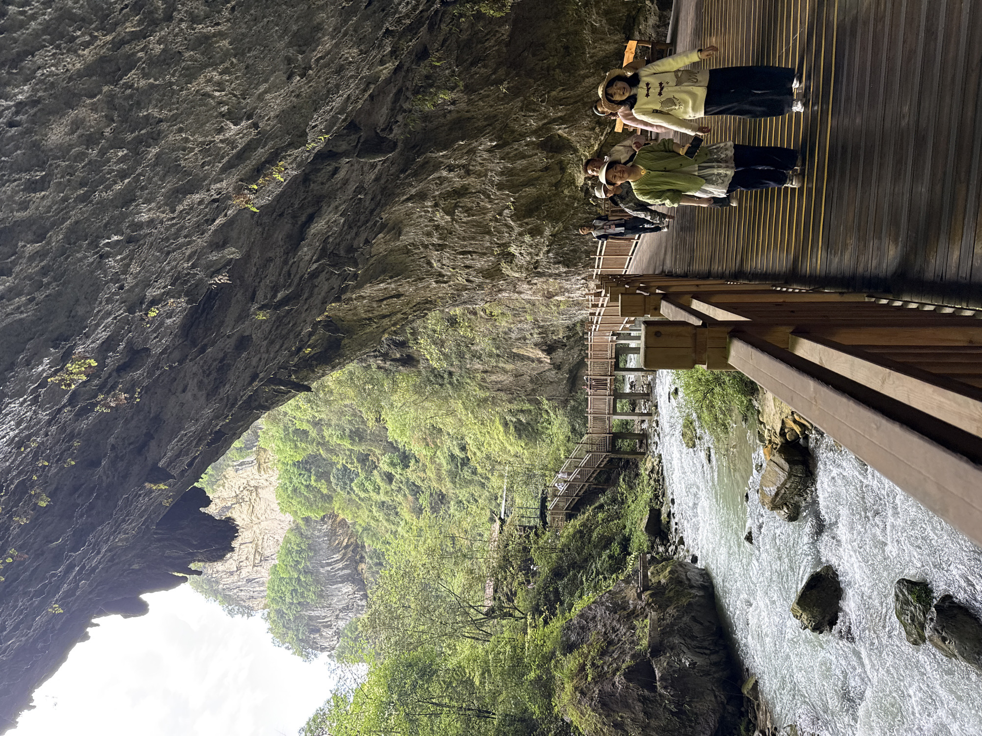 The image shows a view from within a cave, looking out towards a river. The location is in Zhangjiajie, China. A group of people is standing on a wooden walkway inside the cave. The cave ceiling is visible on the left side of the image. A river runs along the right side of the walkway. Lush greenery covers the canyon walls across from the cave entrance.