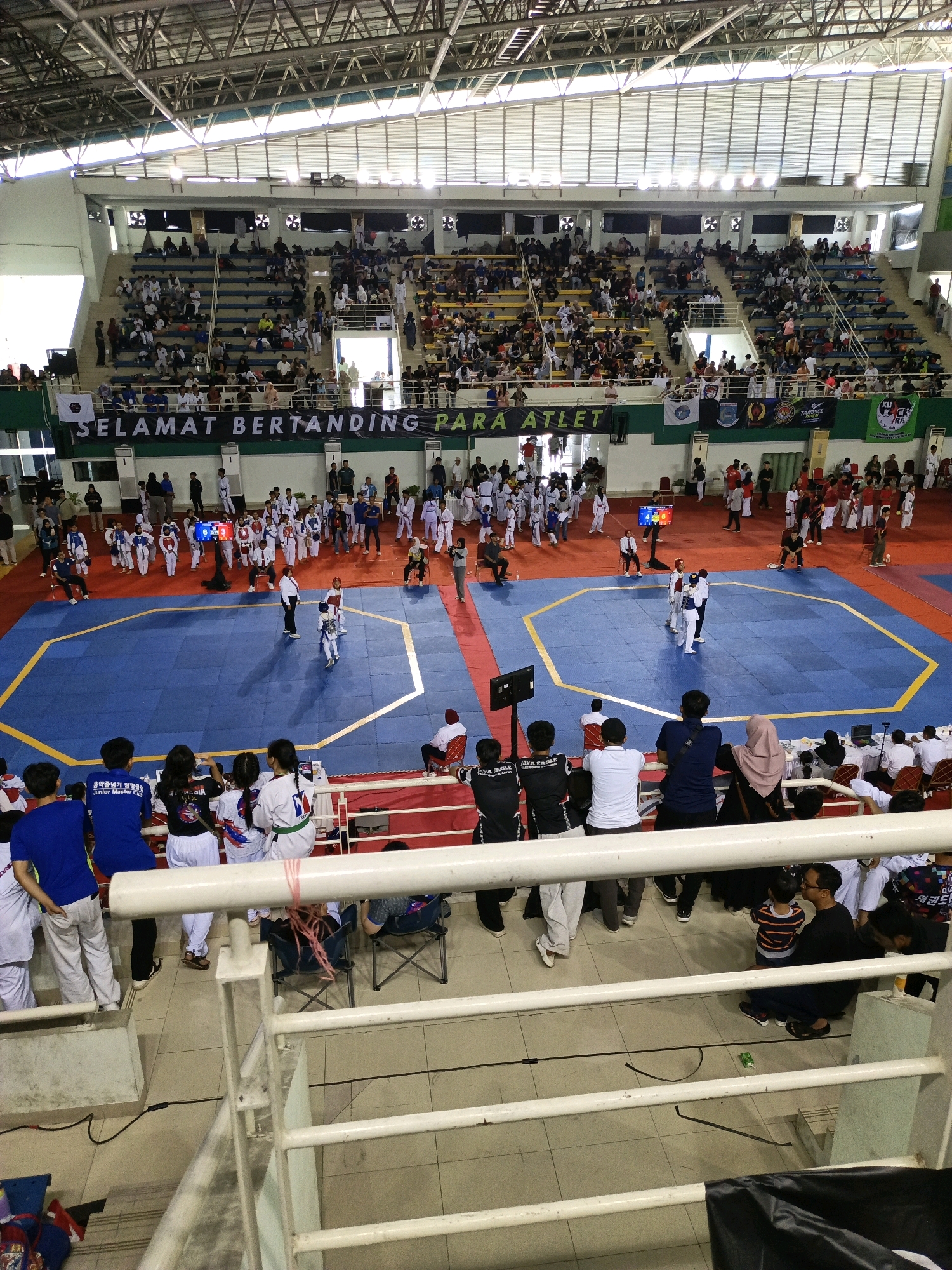 The image captures a Taekwondo competition held indoors at the Convention Center, BSD City, South Tangerang, Indonesia. The venue features a large blue competition mat with yellow boundary lines, surrounded by a red area. Spectators are seated in tiered bleachers on either side of the mat. Several individuals, likely competitors, are dressed in white and black Taekwondo uniforms. Some are actively engaged in matches on the mat, while others are gathered around the edges, waiting their turn or observing the events. The environment is brightly lit, with metal scaffolding and a high ceiling visible. A banner with Indonesian text, "SELAMAT BERTANDING PARA ATLET," which translates to "GOOD LUCK ATHLETES," is prominently displayed above the spectators.