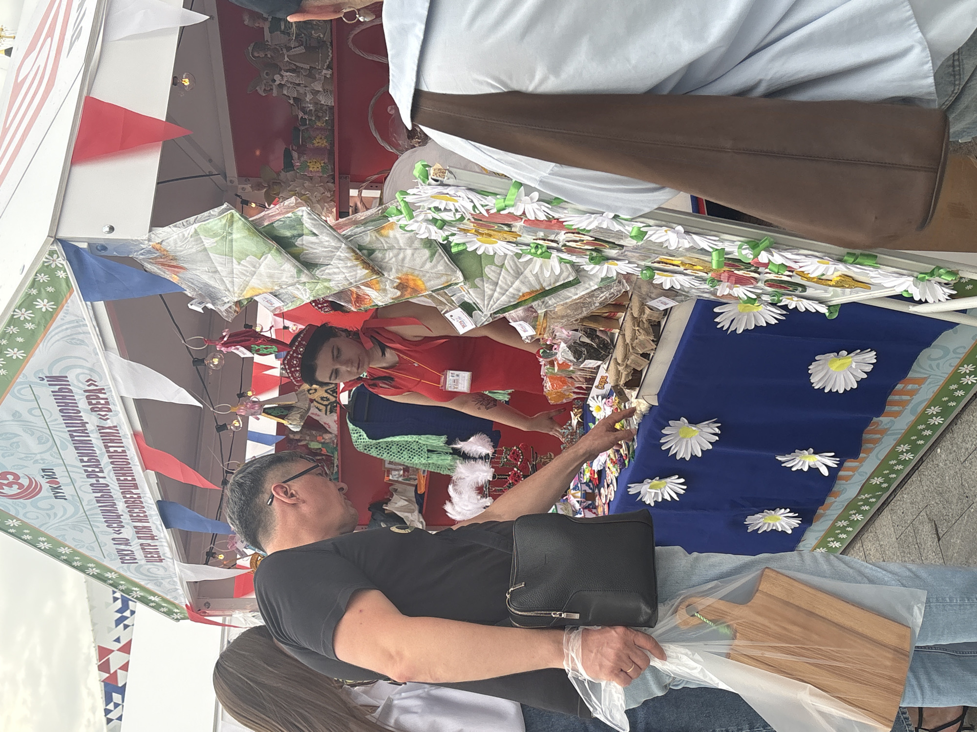 This image captures a candid moment at an outdoor market or fair, likely in Astrakhan, Russia. The scene is vibrant with colorful triangular flags adorning a stall, suggesting a festive atmosphere. The primary focus is on a group of people interacting with merchandise displayed on the stall.
In the foreground, a man wearing glasses and a black t-shirt is seen from the side, holding a black leather bag with a zipper. He appears to be engaged in a conversation or transaction. To his right, a wooden cutting board, wrapped in clear plastic, is visible, indicating the type of goods being sold.
Behind the man, a woman in a red top and a headscarf is partially visible, leaning towards the stall. She seems to be part of the vendor team or a customer. The stall itself is laden with various items, including what appear to be decorative fabric squares with floral patterns, possibly napkins or placemats, and a garland of white daisy-like decorations. A blue fabric with white daisy appliques is prominently displayed.
Above the stall, a banner is visible with Russian text that reads: "ЛУКОЙЛ ГСКУ ДО «СОЦИАЛЬНО-РЕАБИЛИТАЦИОННЫЙ ЦЕНТР ДЛЯ НЕСОВЕРШЕННОЛЕТНИХ «ВЕРА»". This text identifies the stall as belonging to the "Lukoil Social-Rehabilitation Center for Minors 'Vera'". The presence of the Lukoil logo suggests a connection to the oil company.
The lighting indicates it is daytime, possibly late morning or early afternoon, and the weather appears to be fair. The overall impression is one of a community event or a local market, with a focus on handmade crafts or local products. The people's expressions are difficult to discern clearly due to the angle and resolution, but the general atmosphere seems relaxed and engaging.