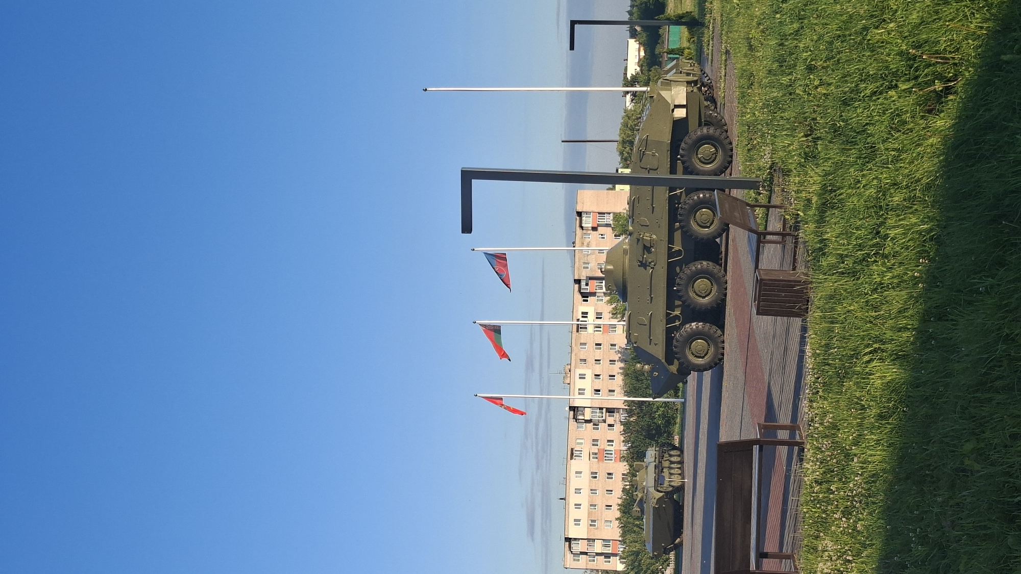 The image captures a sunny outdoor scene in the city of Smolevichi, Belarus, featuring a memorial or display of military vehicles. In the foreground, lush green grass dominates the lower portion of the frame, with sunlight casting shadows. Two wooden benches and a trash receptacle are placed on a paved walkway. Behind the seating area, two military armored vehicles are displayed. The larger vehicle, an eight-wheeled armored personnel carrier, is prominently positioned. Further back, a tracked armored vehicle is visible.
Three flags are hoisted on white poles behind the military vehicles. The flags appear to be red, green and black, red and black, and red, white and green. A multi-story apartment building with numerous windows stands in the background, suggesting an urban or semi-urban setting. The sky is clear and blue, indicating fair weather. Two modern-style lamp posts are also visible within the scene. The overall impression is that of a public space, possibly a park or town square, dedicated to military history.