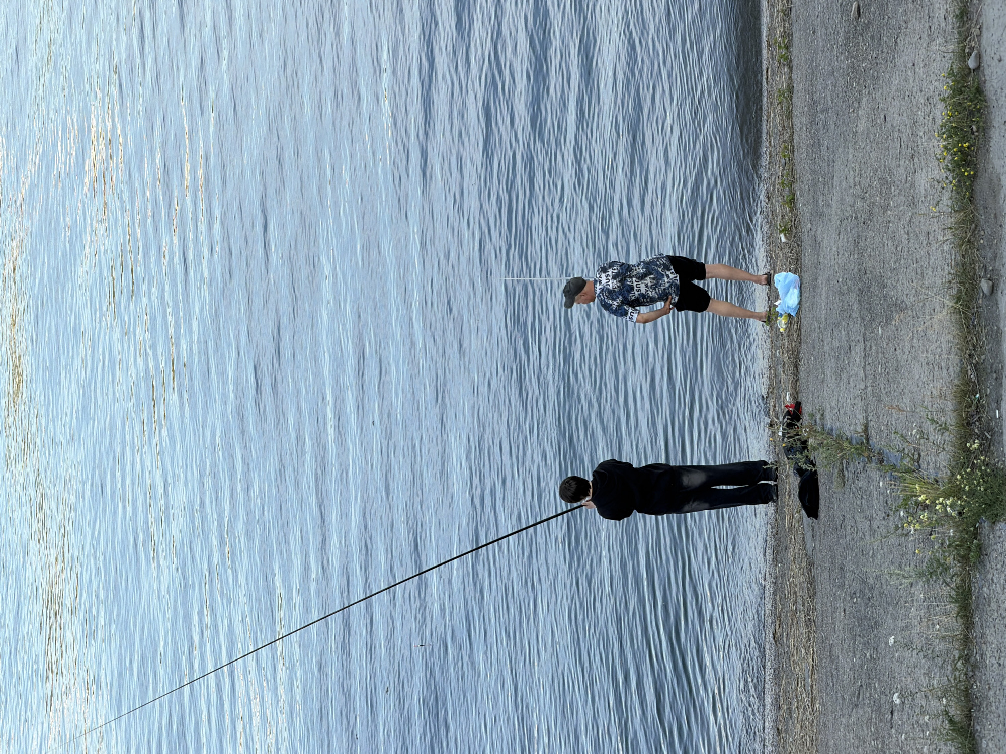 Two individuals are fishing on the bank of a body of water. The person on the left, facing away from the camera, is wearing a black hoodie and dark pants. They are holding a long fishing rod that extends out over the water. The person on the right, also facing away from the camera, is wearing a patterned t-shirt, black shorts, and a baseball cap. They are also holding a fishing rod, which is shorter than the other. In the foreground, on the concrete embankment, there are a few items including a black bag, a red object, and a blue plastic bag. The water is calm with ripples, reflecting the light. The environment appears to be a riverbank or lake shore. This scene could be located along the Angara River in Irkutsk, Russia, given the typical urban waterfront environments there.