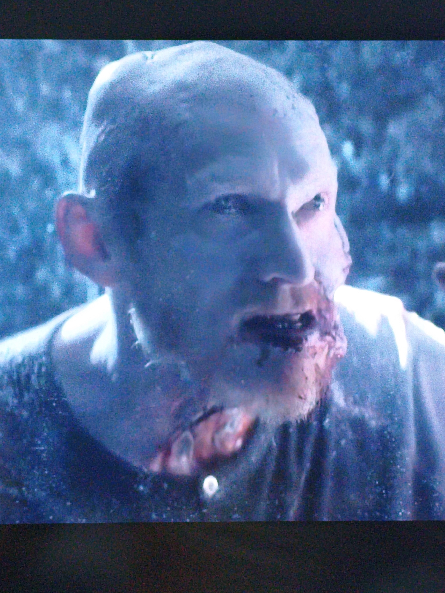 This is a close-up, grainy, and darkly lit image of a person's face, appearing to be from a video or screenshot. The person is bald and has a pale complexion. Their head is tilted slightly to the left. There is significant gore around the mouth and jaw area, with blood and what appears to be torn flesh visible. The background is blurred and out of focus, with a predominantly dark blue and purple hue, suggesting an outdoor or dimly lit environment. There are some faint, lighter specks scattered throughout the background, possibly indicating water droplets or dust. The lighting is harsh and casts strong shadows, particularly on the right side of the face. The image does not contain any recognizable landmarks or elements that would allow for identification of a specific location within Vladivostok, Russia.