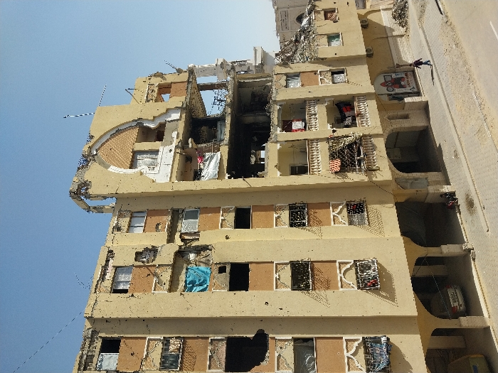 An exterior daylight photograph depicts a multi-story building exhibiting extensive damage in Khan Yunis, Palestinian Territory. The structure, appearing to be a residential or mixed-use building of light brown/beige stucco with reddish-brown accents and ornate window grates, shows significant structural compromise. Large sections of the upper right and central facades are destroyed, exposing interior rooms, collapsed floors, and twisted rebar. Soot and burn marks are visible on the middle floors. Numerous windows are broken or missing, with one on the left covered by a blue tarp. Bullet or shrapnel impacts mark the building's exterior.
On the ground level, a series of arches form a recessed walkway or parking area. A white sedan is parked under one arch on the left. Two individuals are seated on the sidewalk under an archway on the left. Further to the right, a single individual walks on the paved sidewalk from right to left, passing a partially visible storefront sign with a red image and Arabic text. Damaged balconies with decorative balustrades have clothing hanging from them.
In the background to the right, additional structures appear heavily damaged or reduced to rubble. A clear blue sky is visible above the building.