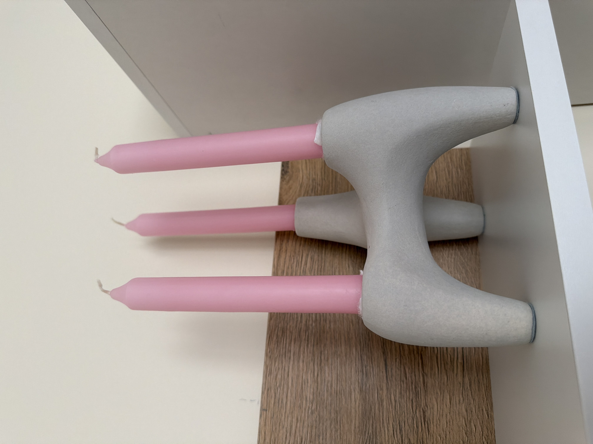 A close-up, eye-level, indoor shot captures a light gray, abstract-shaped concrete candleholder with three pink, tapered candles inserted into it. The candleholder has a unique, organic form with rounded legs and a central structure that holds the candles. The background consists of a textured wooden panel with a light beige wall above it. The scene is set on a white shelf. The image is well-lit, with soft shadows, and appears to be taken in a domestic environment. There are no people present in the image. The location of Pazardzhik, Bulgaria is not discernible from the visual information provided.