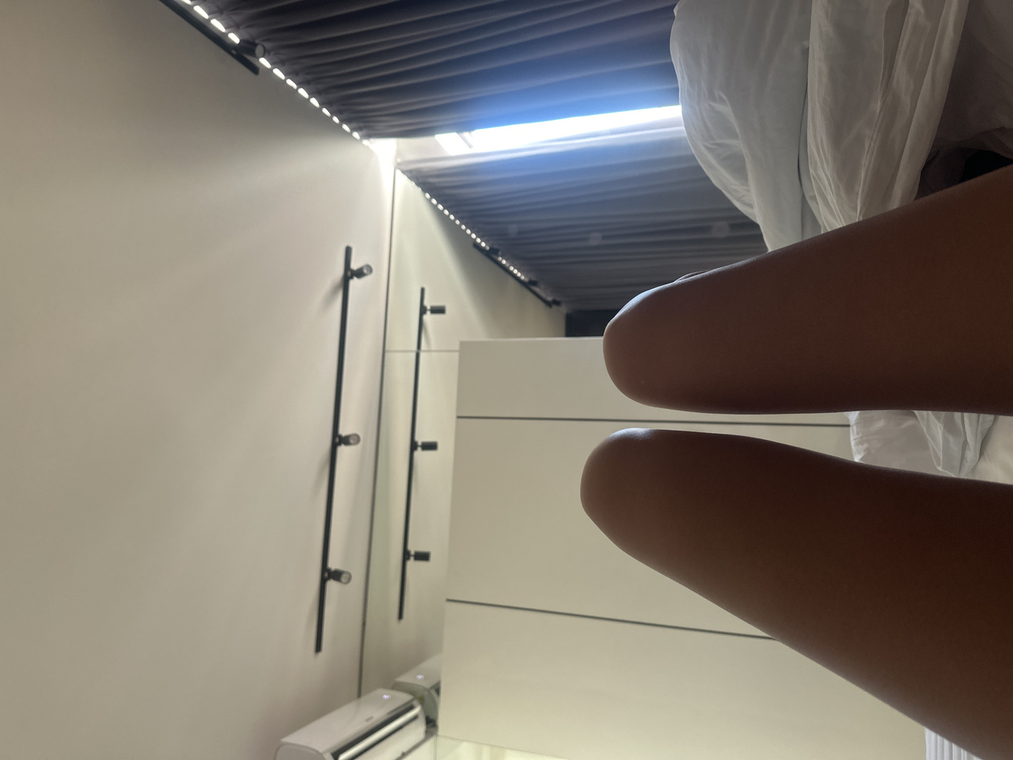 A first-person perspective shot shows two legs with dark skin resting on a white duvet in a bedroom. Above the legs, a white wall and ceiling are visible. A white air conditioning unit is mounted on the wall to the left. Two black track lights with spotlights are attached to the ceiling. To the right, dark gray curtains are drawn, with a bright light filtering in from a window behind them. The room appears to be in North Kuta, Indonesia, suggesting a hotel room or residential bedroom.