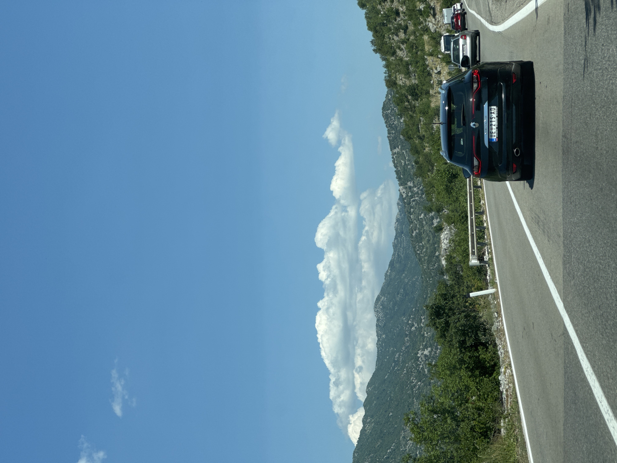 The image depicts a view from a vehicle traveling on a highway in the mountainous region of Cerovo, Montenegro. The road curves to the right, with a white line marking the lane division. A dark-colored Renault Megane station wagon, bearing the license plate "UE 215 AD," is the primary subject in the foreground, traveling in the same direction. Several other vehicles are visible ahead, including a white van, a silver sedan, and a red SUV. The landscape consists of lush green mountains covered in trees and vegetation, with rocky outcrops visible. The sky is a clear blue with scattered white clouds. A metal guardrail lines the left side of the road, separating it from the steep, vegetated embankment.