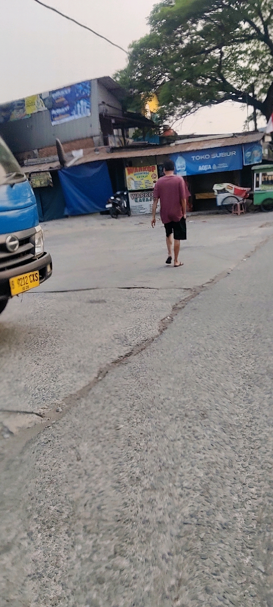In Mekarmukti, Indonesia, a man walks away from a blue vehicle on a street with a cracked asphalt surface. The man wears a maroon t-shirt and black shorts. Behind him, a building with several signs is visible. One sign, partially obscured, reads "WARKOP". Another sign displays "TOKO SUBUR AQUA". The scene is outdoors, with a tree overhead and what appears to be a cart with red and white accents. The sky is overcast.