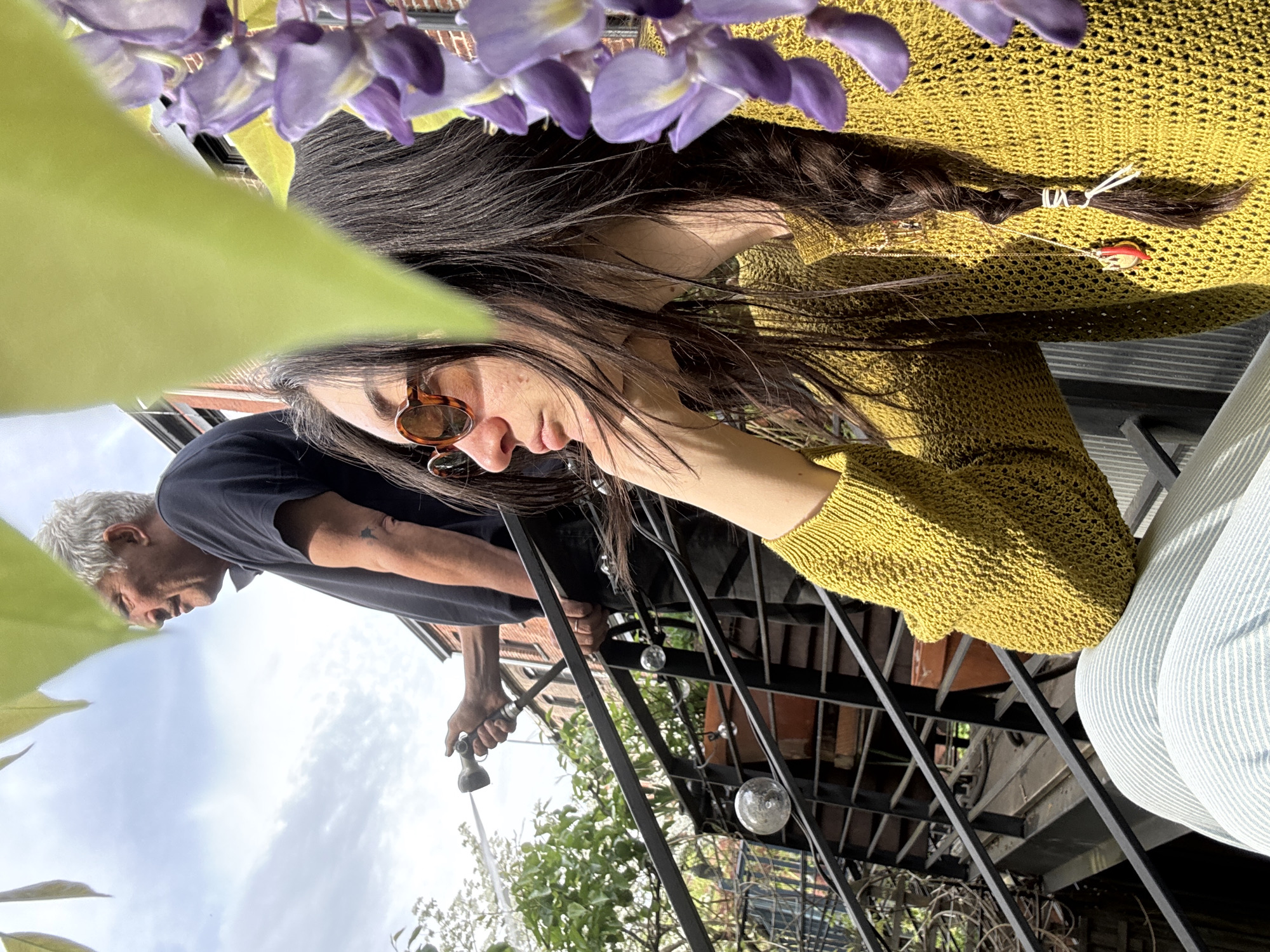 The image appears to show a woman in a mustard yellow sweater, wearing sunglasses, sitting on a metal staircase outdoors. She has long, dark hair, partially braided and secured with a white band. Her face is tilted downward.
A man is visible in the background, appearing to water plants with a hose. He is wearing a dark shirt and has gray hair.
The location is likely an outdoor patio or fire escape in an urban setting, possibly New York City, United States of America, due to the presence of brick buildings, metal staircases, and urban greenery. Purple wisteria flowers are hanging overhead, obscuring part of the image. There are also lightbulb decorations on the stairs.