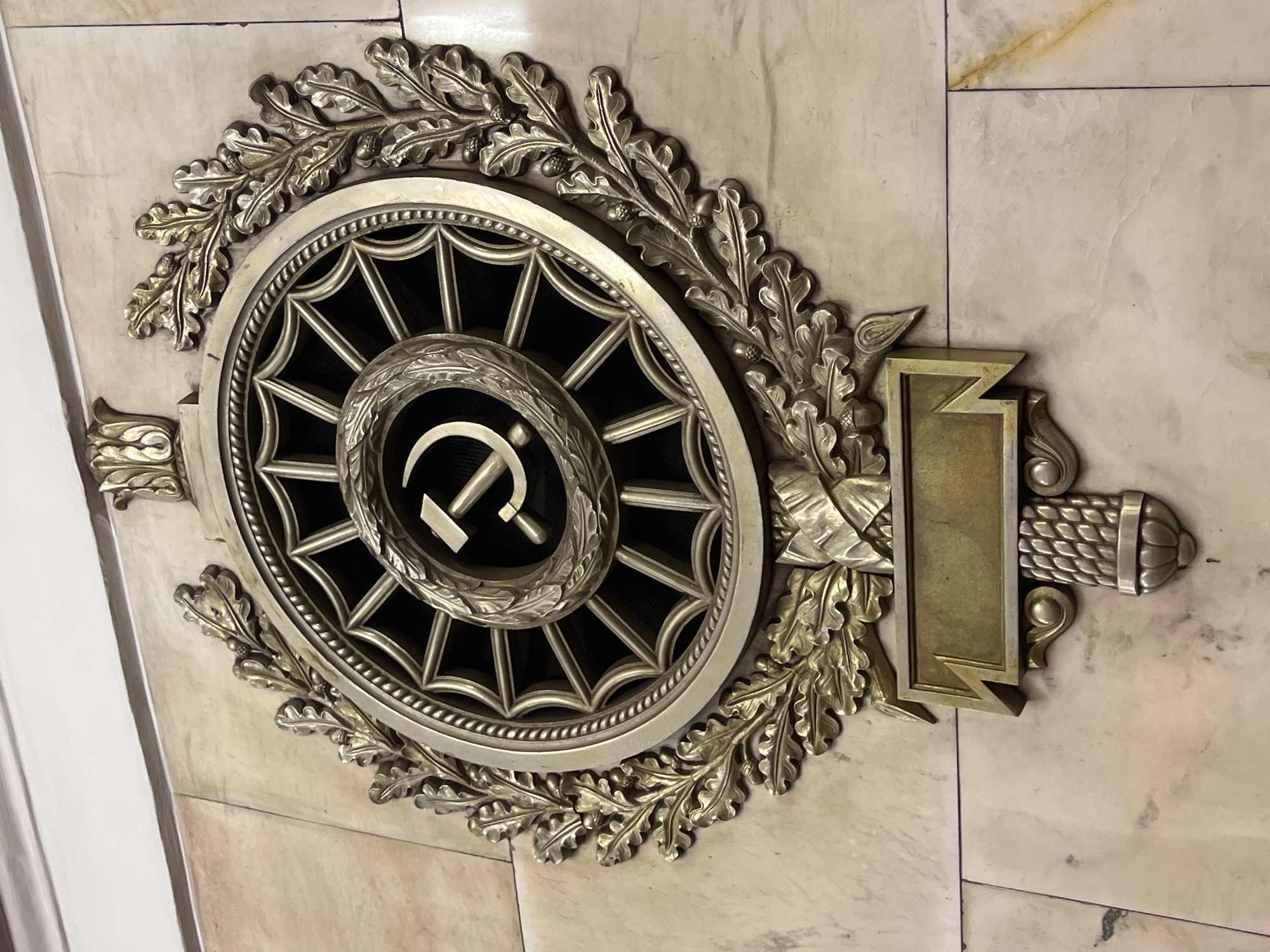 The image features an ornate, bronze-colored decorative element mounted on a marble-like wall. The centerpiece is a circular grille with a starburst pattern. In the center of the grille is the hammer and sickle emblem. The grille is surrounded by a wreath of oak leaves and acorns. To the right of the circular design is a rectangular plaque-like element with decorative detailing. The image appears to be of an architectural detail likely found in a building in Saint Petersburg, Russia.